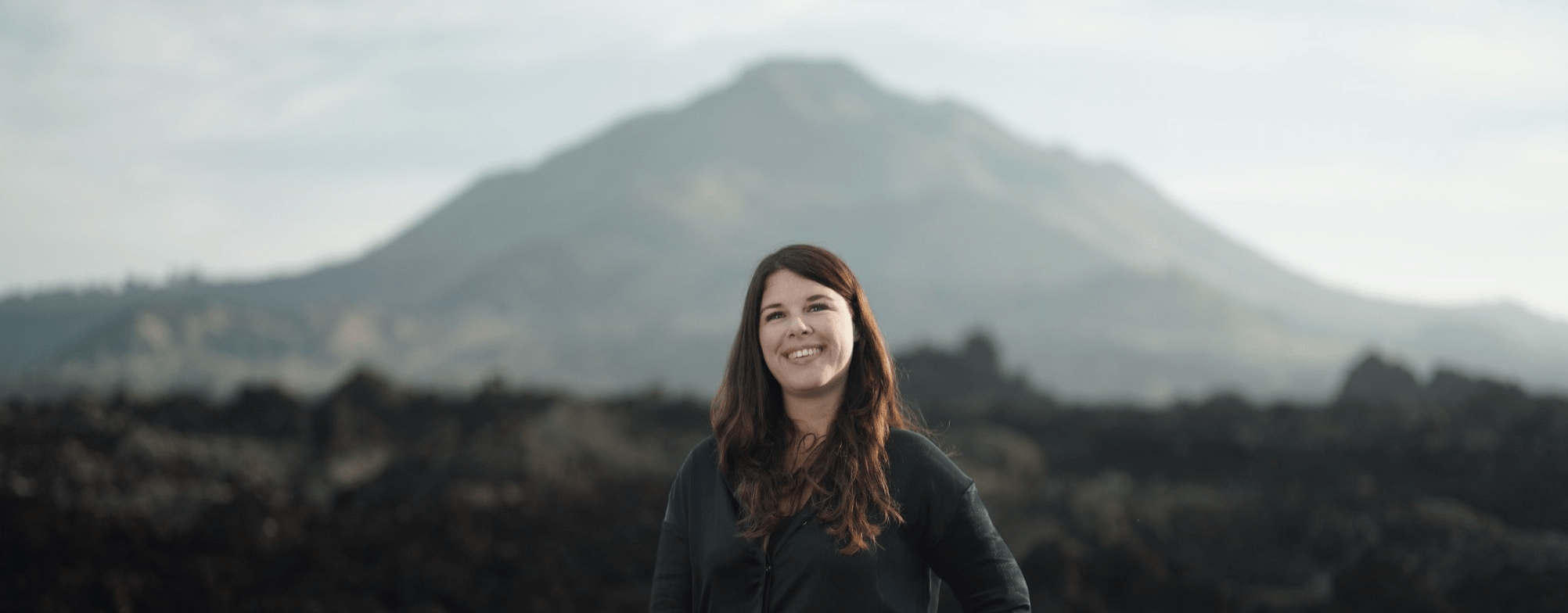 A woman with long reddish-brown hair smiling outdoors with a volcano in the background.