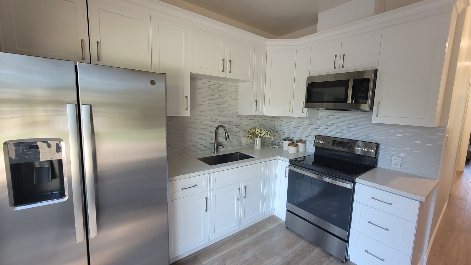 Modern kitchen with white cabinets, stainless steel refrigerator, stove, microwave, sink, and light wood flooring.