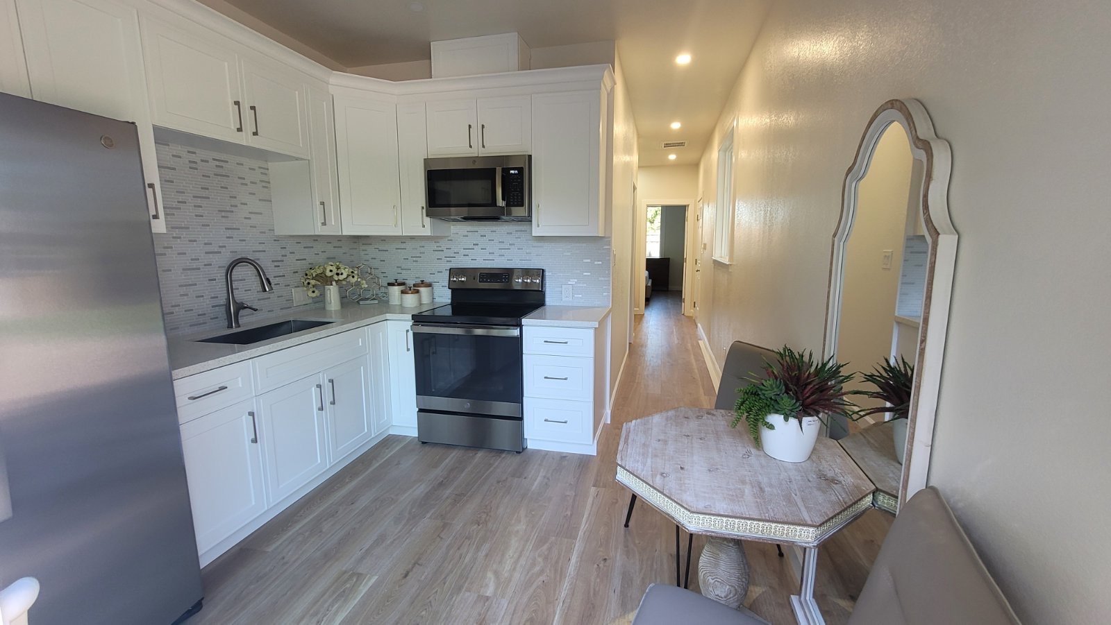 Modern kitchen with white cabinets, stainless steel appliances, a small dining table with chairs, and decorative plants. Light wood flooring and tile backsplash detail the space.