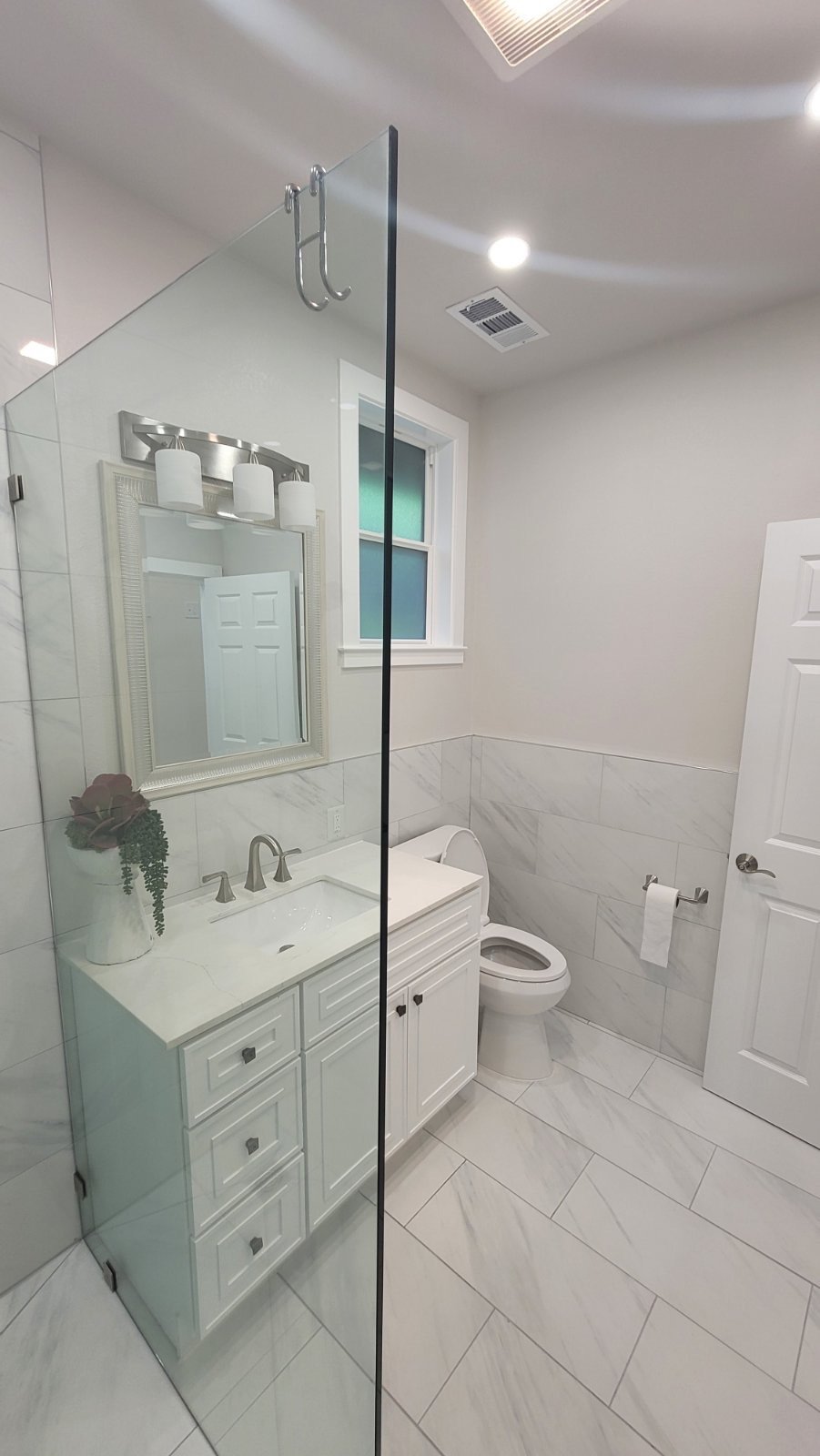 Modern bathroom with glass shower, white vanity, sink with faucet, toilet, and wall-mounted mirror. Light gray tiles and a small window enhance the space.