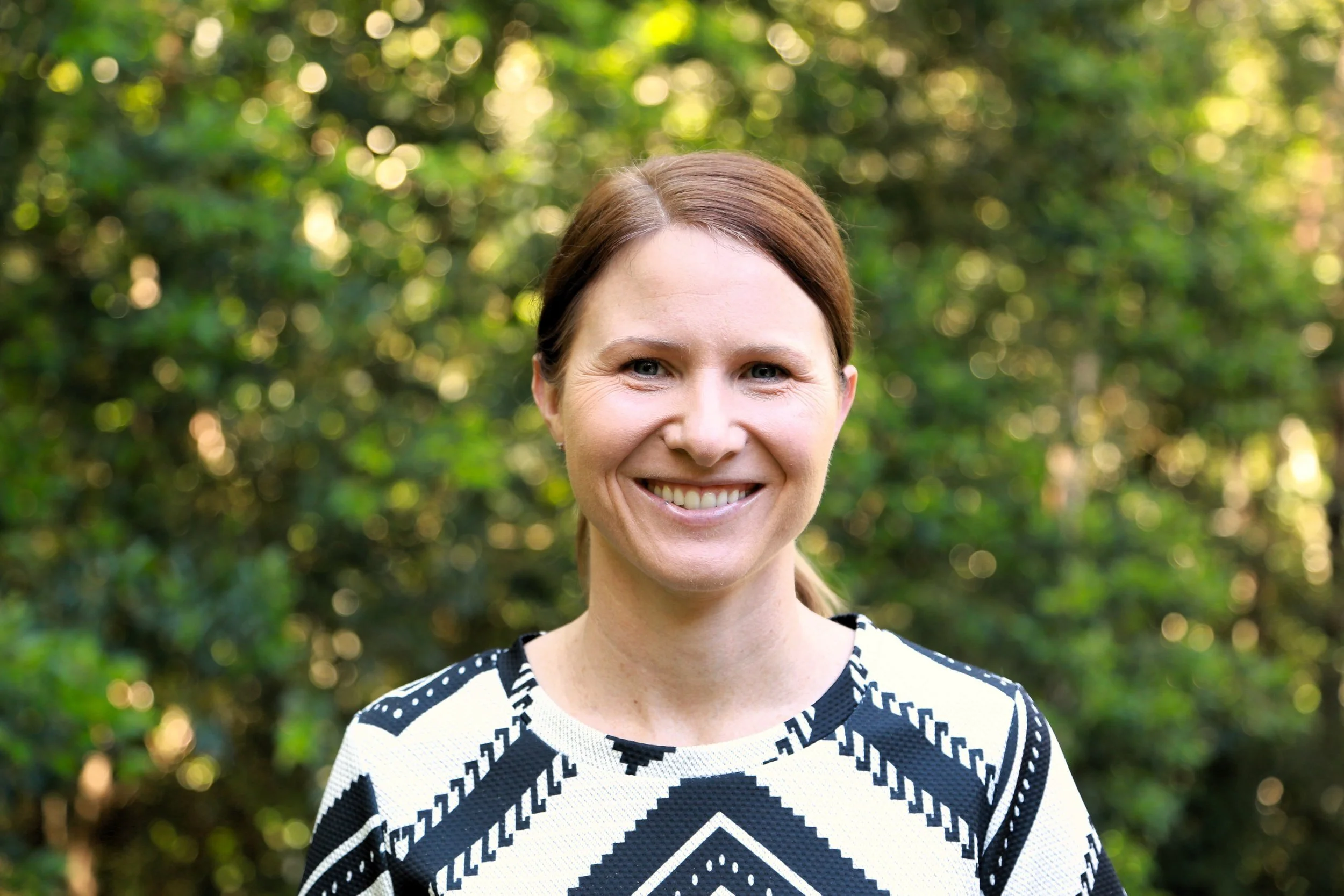 A woman with red hair and blue eyes smiling outdoors in front of green foliage, wearing a black and white patterned sweater.