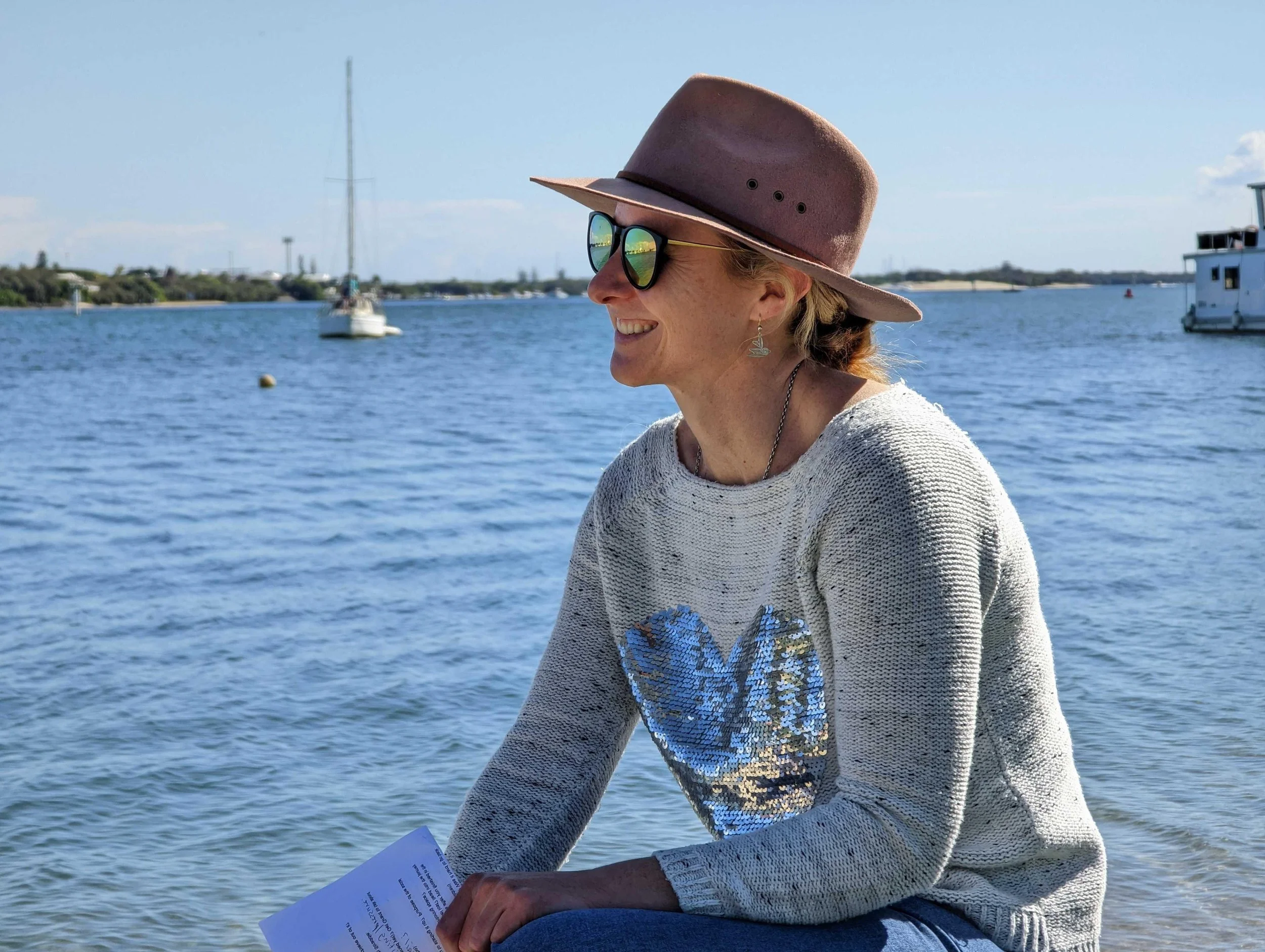 A woman with blonde hair wearing a wide-brimmed brown hat, sunglasses, and a gray sweater with sequins, sitting by the water when smiling. A sailboat, a small boat, and other boats are visible in the background on a sunny day.