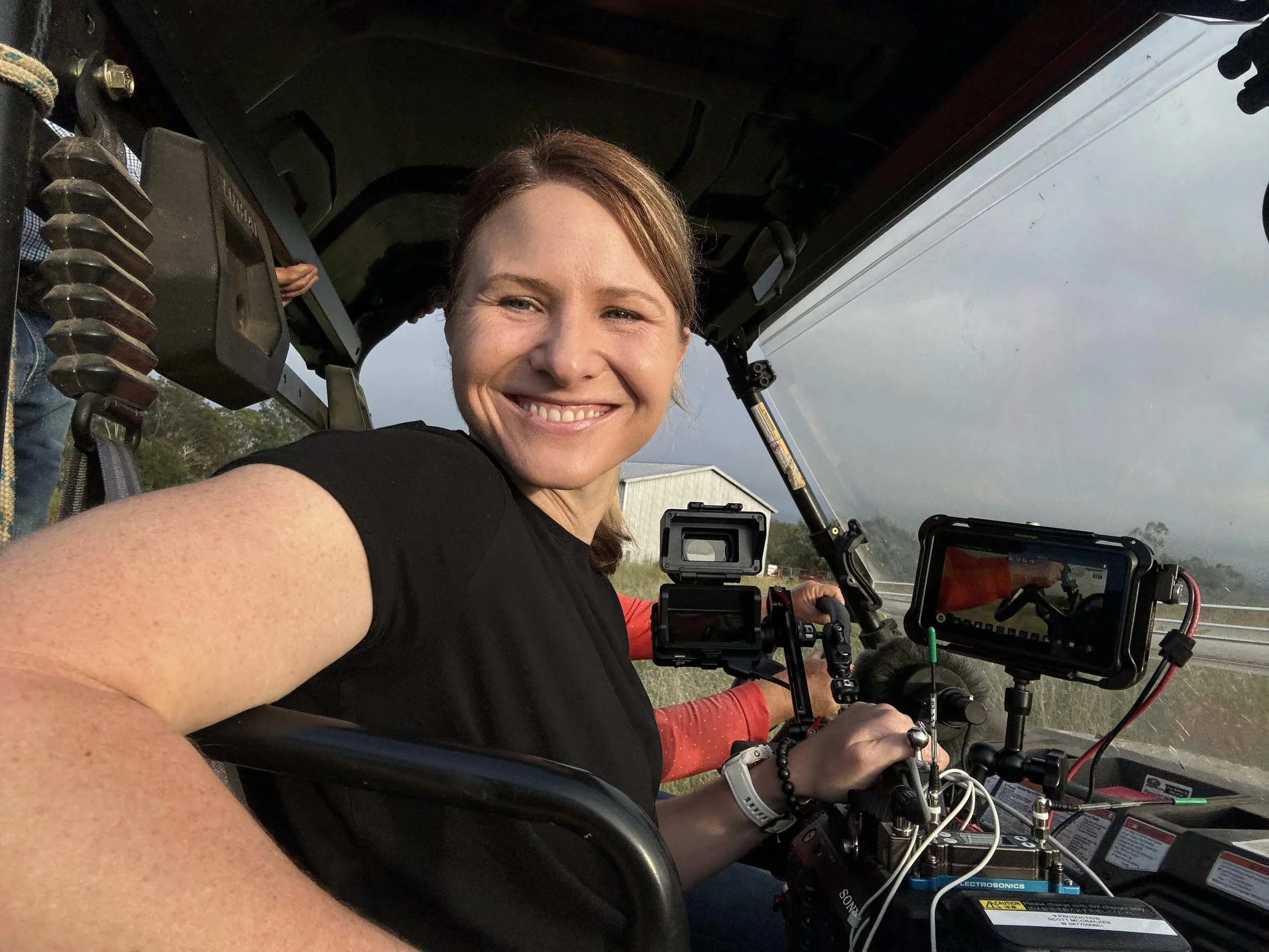 A woman with brown hair is smiling and taking a selfie inside a cockpit or control area of an aircraft or helicopter, with equipment, screens, and controls visible around her.