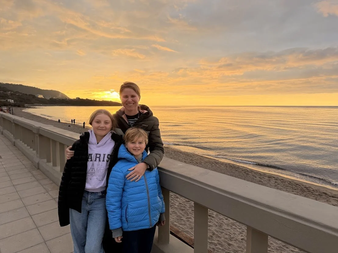 Three people, a woman and two children, standing by a pier railing on a beach at sunset, with calm ocean waters and a colorful sky in the background.