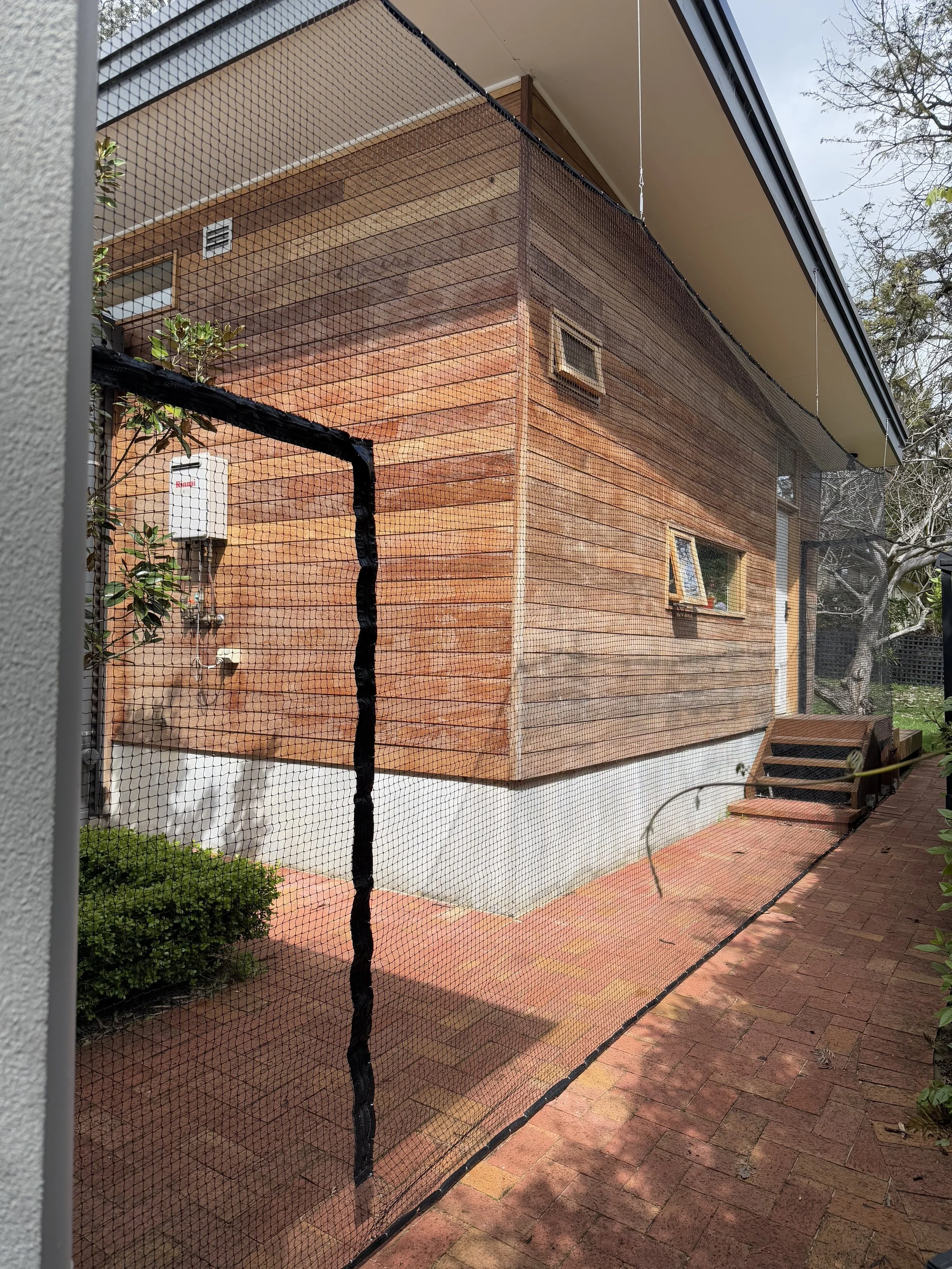 Backyard view of a house with a wooden exterior, small windows, a brick walkway, and a screened-in area with a black net.