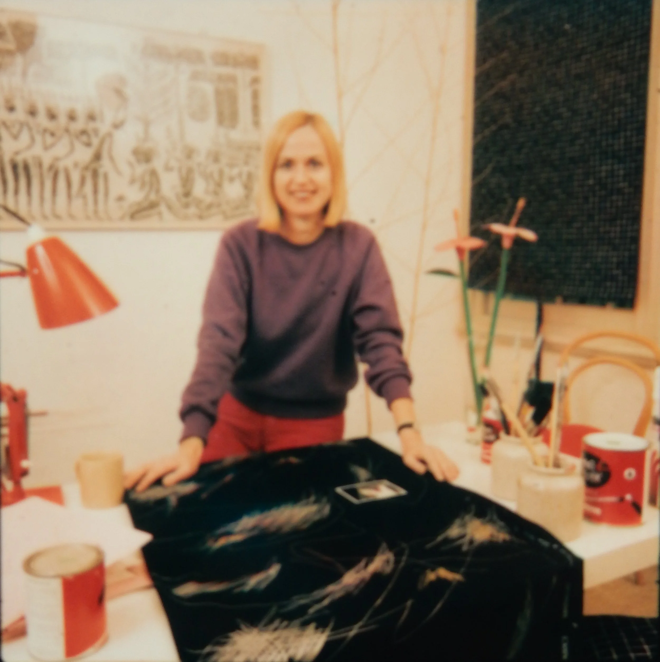 A woman with short blonde hair wearing a purple sweater and red pants is smiling while standing behind a table with art supplies and a large black bag in a room decorated with artwork and plants.