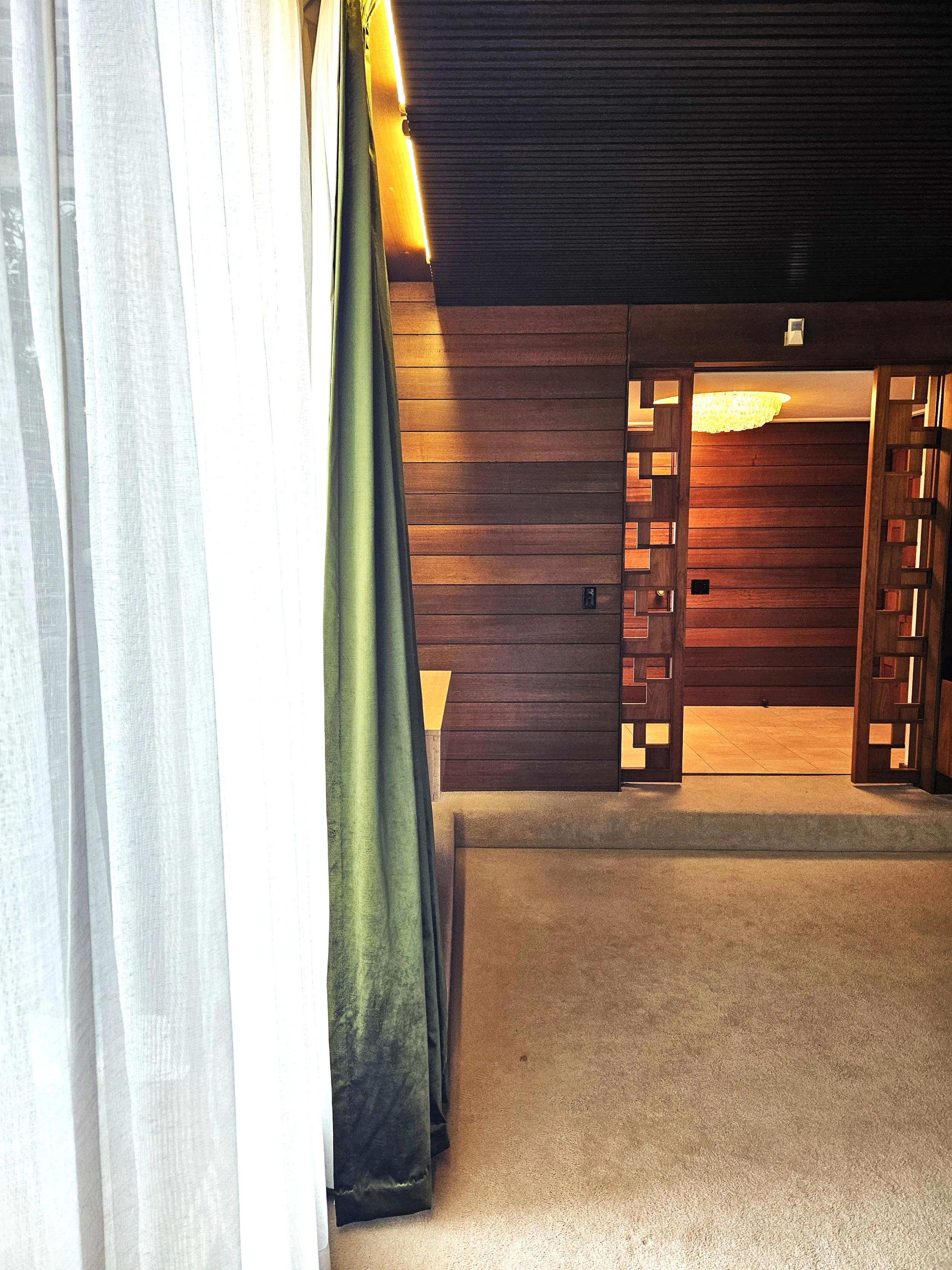 Photo of an interior room with a window and white sheer curtains, a green curtain, a beige carpet, and a wooden partition leading to another room with a tiled floor and a chandelier.