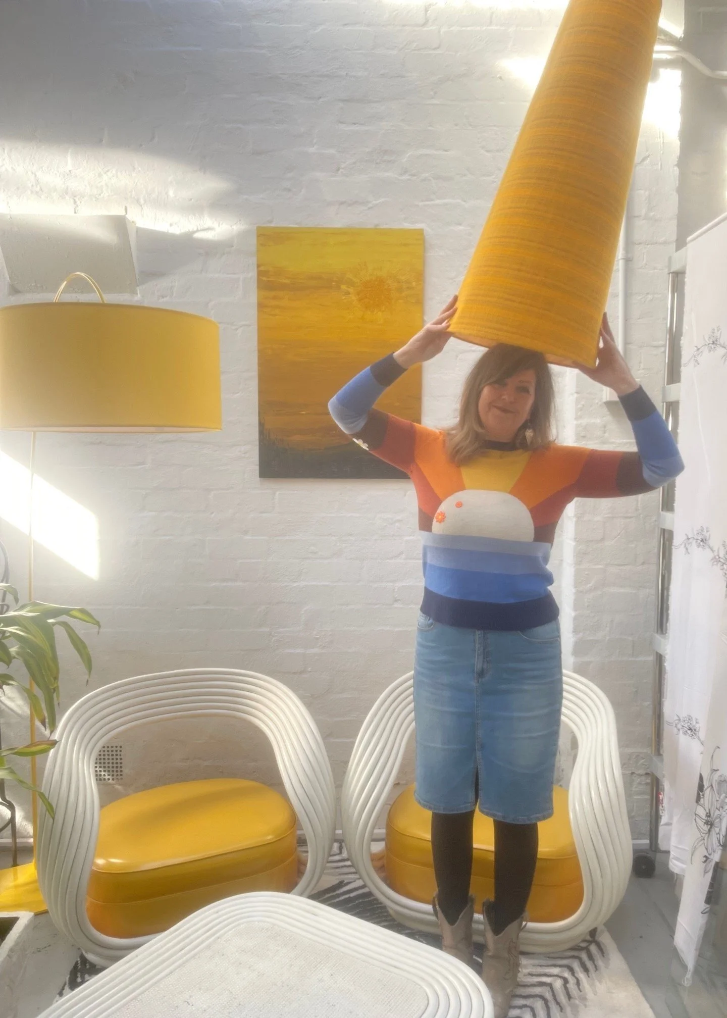 A woman standing indoors, holding a large yellow lampshade above her head. She is wearing a colorful, abstract-patterned sweater, a denim skirt, black tights, and beige boots. The room has white brick walls, yellow and white chairs, a tall yellow lamp, and a piece of artwork on the wall.