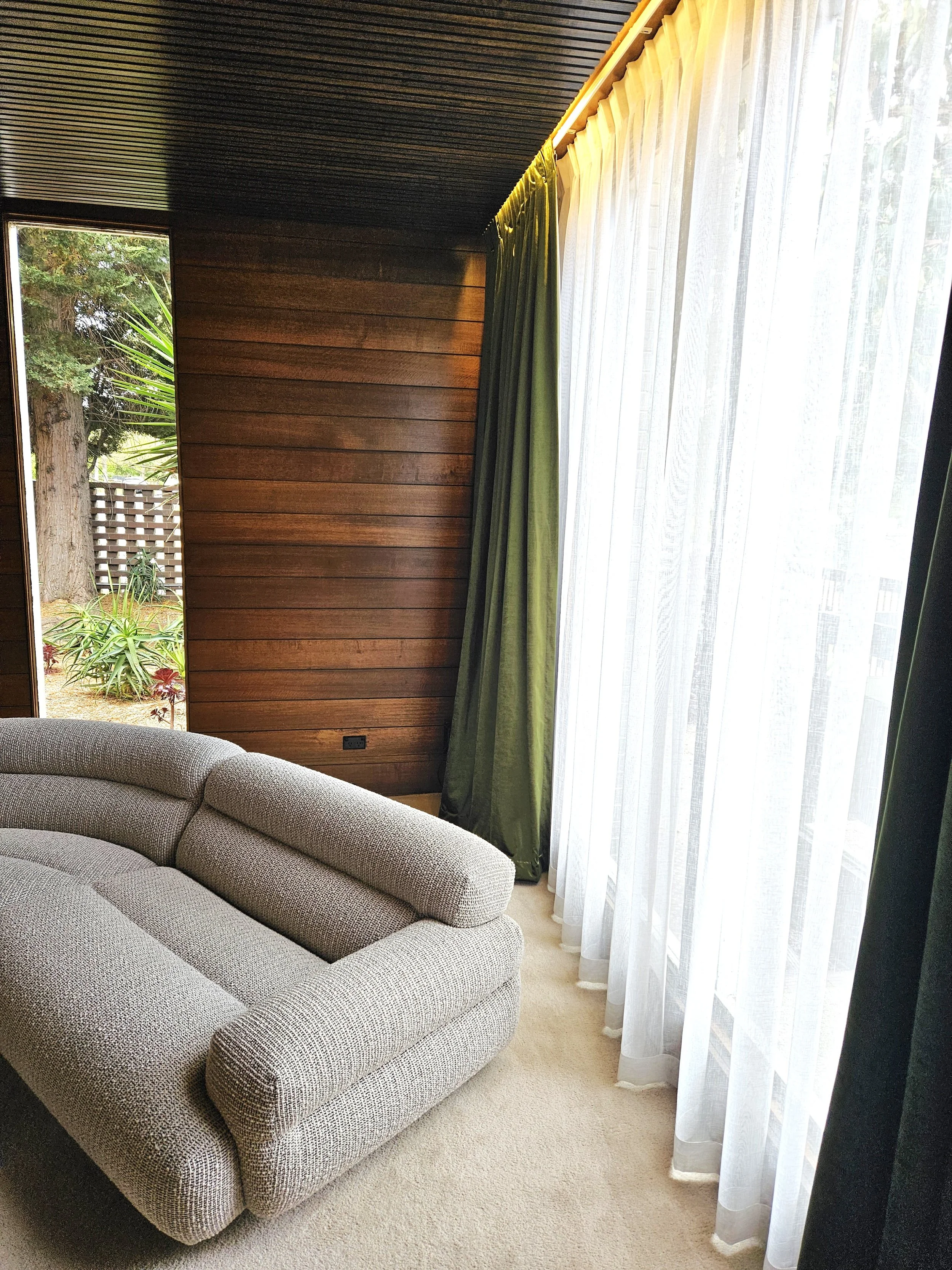 A cozy living room with beige fabric sofa, wooden wall paneling, and large window with white sheer curtains and green drapes, overlooking a garden outside.