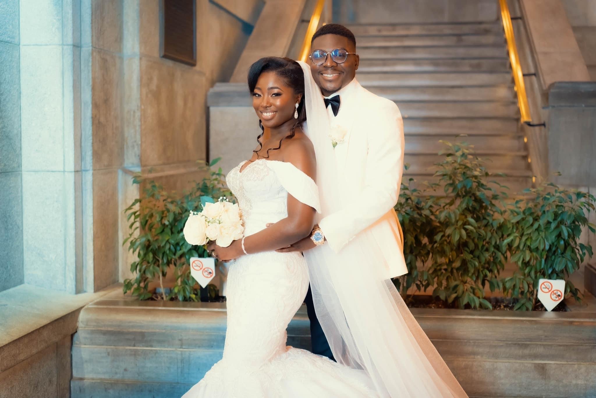 A bride and groom posing on a staircase in elegant wedding attire, with the bride holding a bouquet of white flowers. They are smiling and surrounded by greenery.