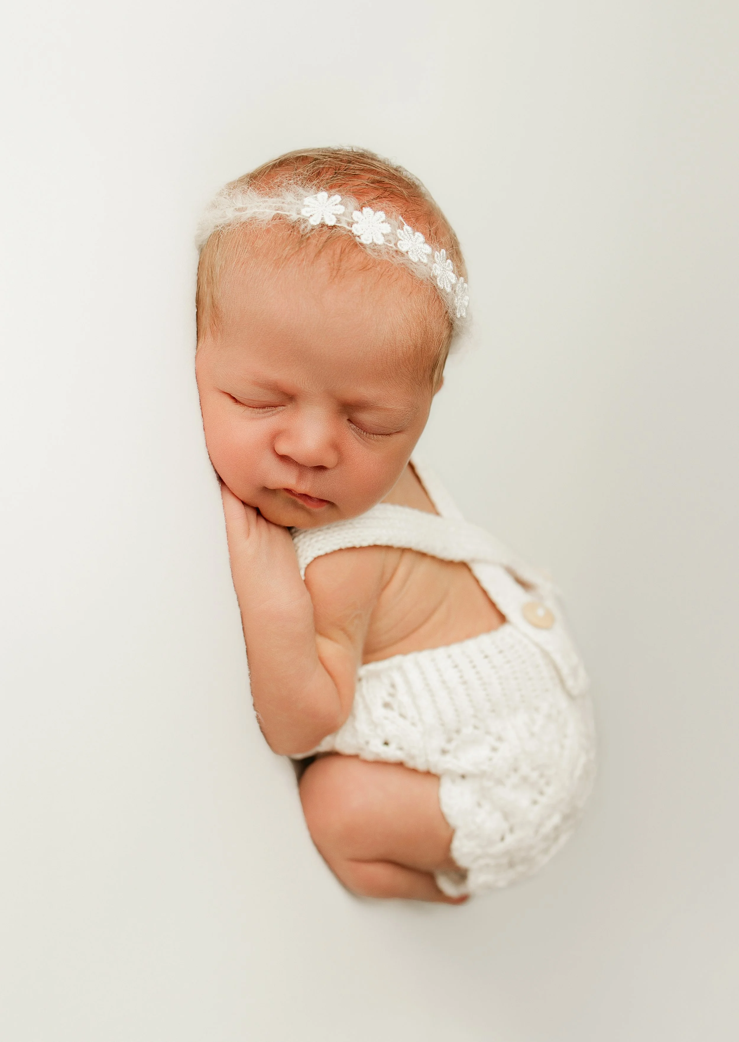 A sleeping newborn baby in a white knitted outfit with a flower headband, resting on a white surface.