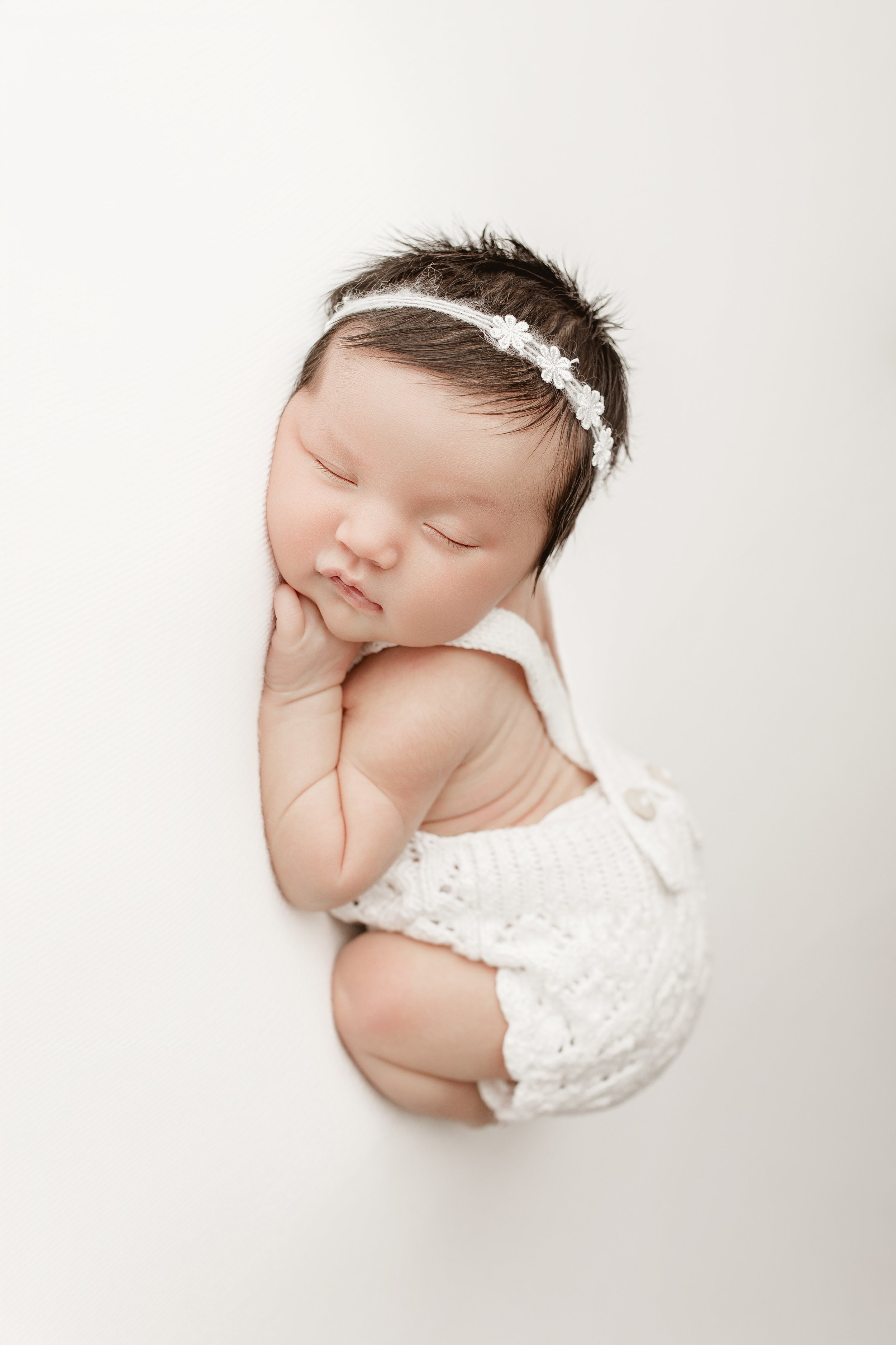 A peaceful newborn baby girl sleeping, resting her head on her arm, wearing a white knitted outfit and a delicate floral headband.