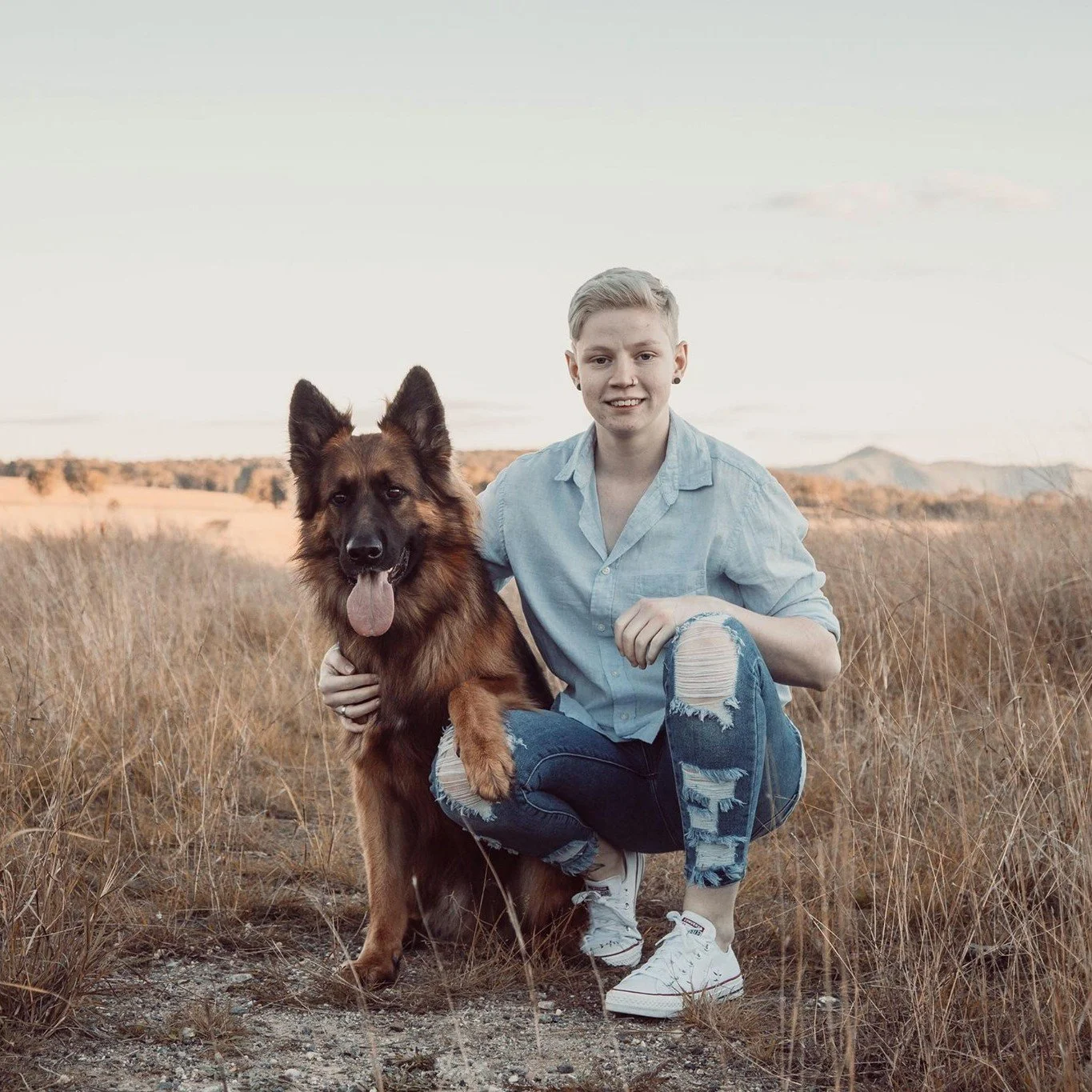 A young person with short blonde hair wearing a light blue shirt and ripped jeans kneeling beside a German Shepherd dog in a grassy field during sunset.