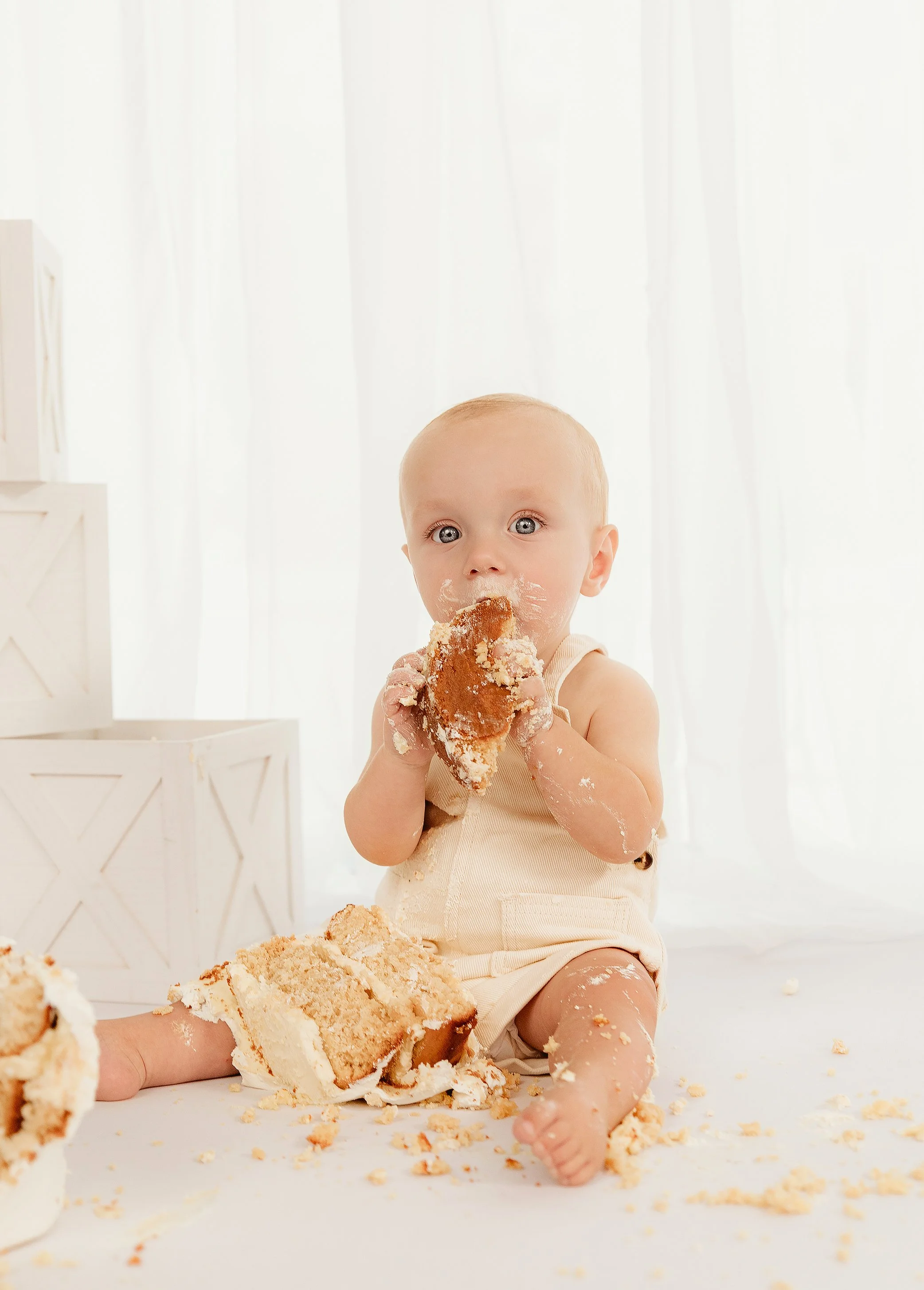 A young baby sitting on a white surface, eating a large slice of cake with frosting, surrounded by cake crumbs and pieces, with a bright white curtain background.