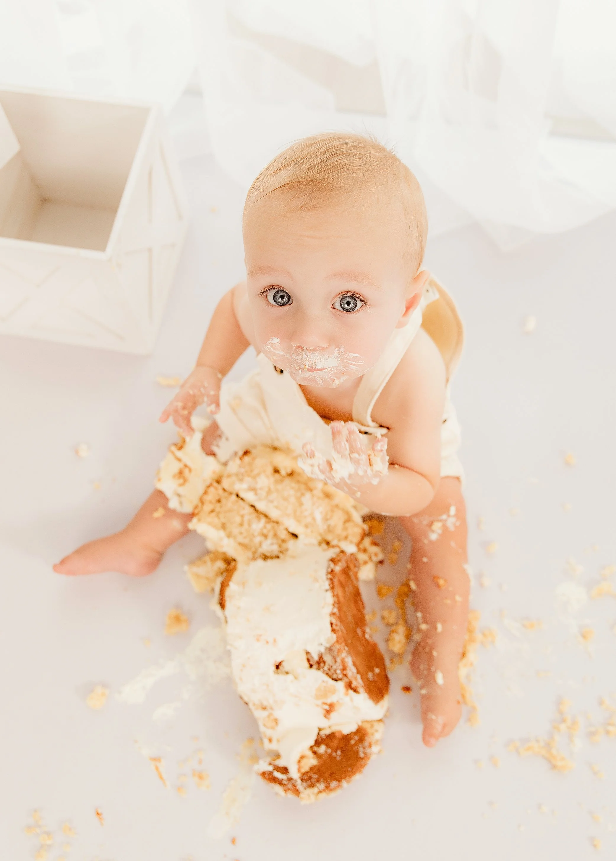 A young child with blonde hair and blue eyes sitting on a white surface, covered in cake and frosting, holding a large piece of cake, with a surprised expression.