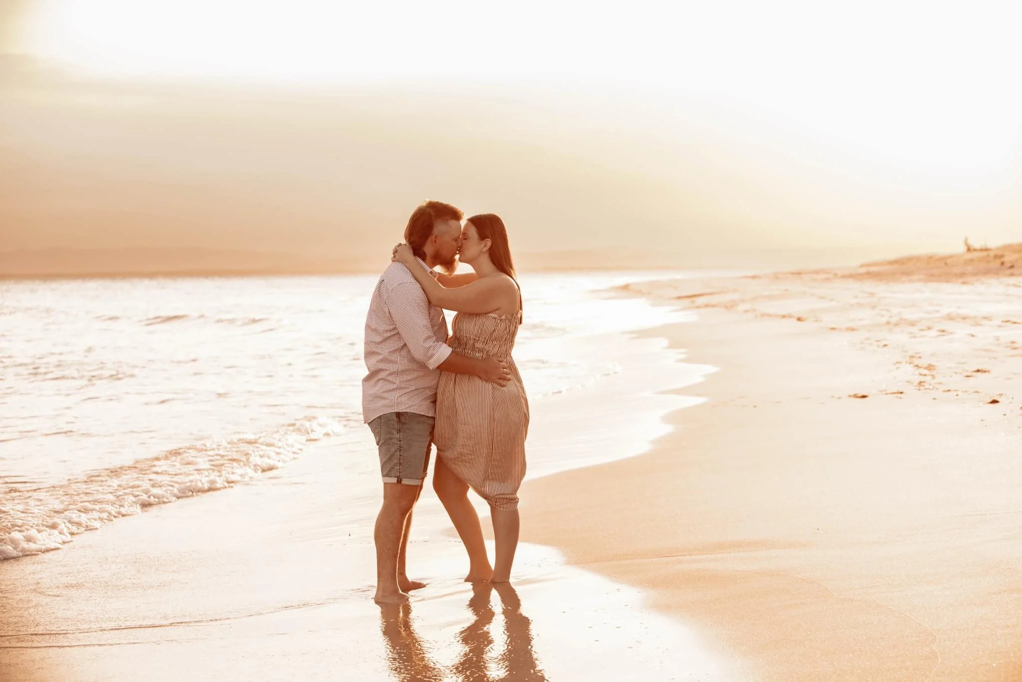 A couple standing in the shallow waves on the beach at sunset, embracing and leaning in for a kiss.