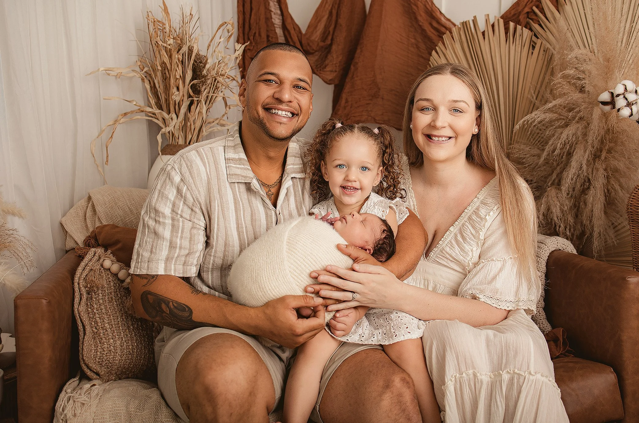 A happy family of four sitting together on a cozy sofa. The father, mother, young girl, and newborn baby are smiling and looking at the camera. The baby is wrapped in a white blanket and appears to be sleeping. The family is surrounded by neutral-col