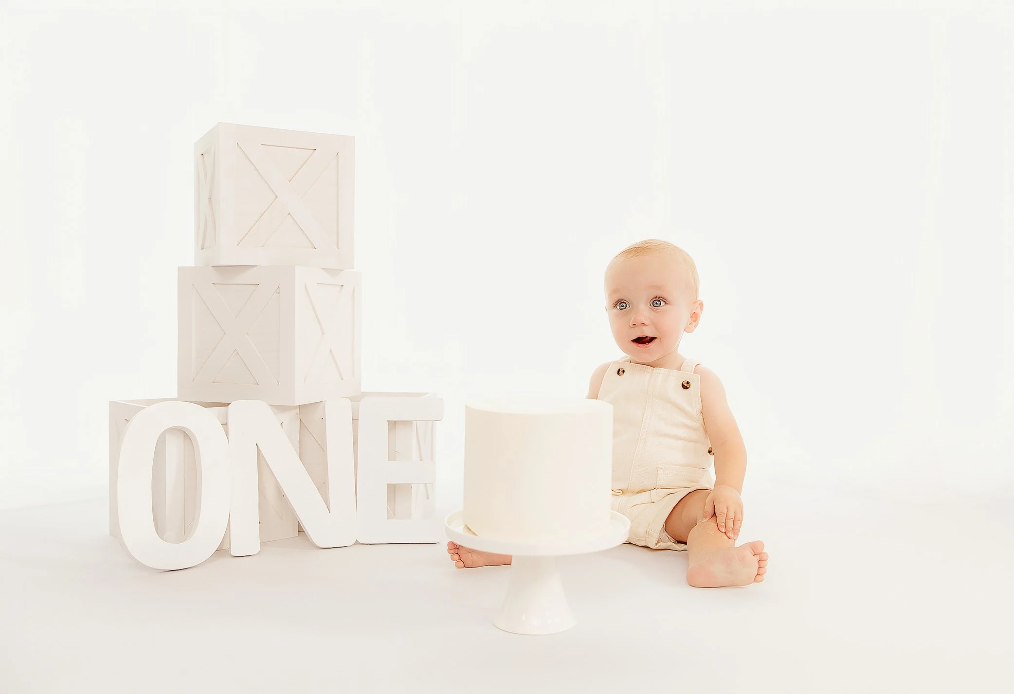 Baby sitting on the floor next to a white cake with a 'LOVE' sign and wooden crates spelling 'ONE' in a bright, minimalist setting.