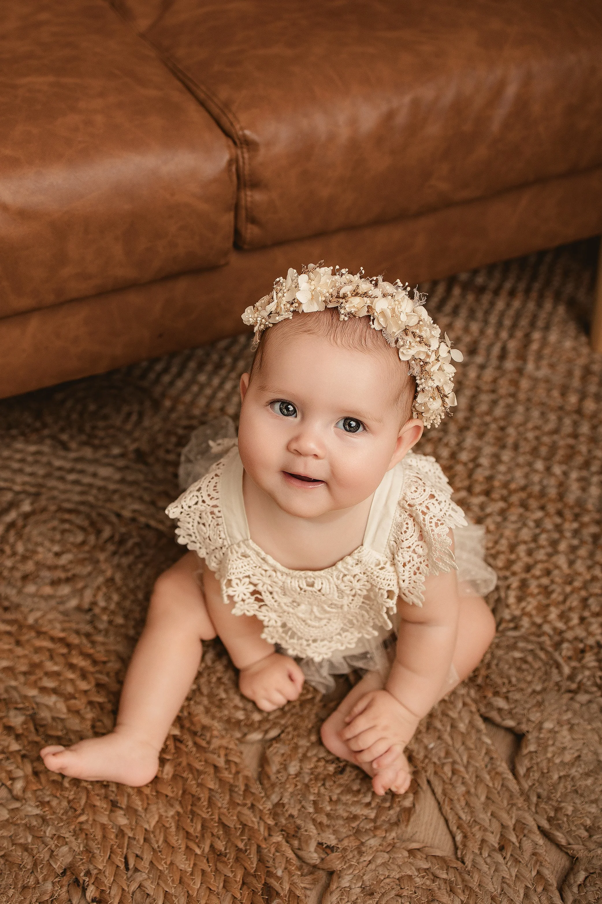 Baby girl sitting on a brown textured rug, wearing a cream lace dress and a cream floral headband, looking up at the camera.