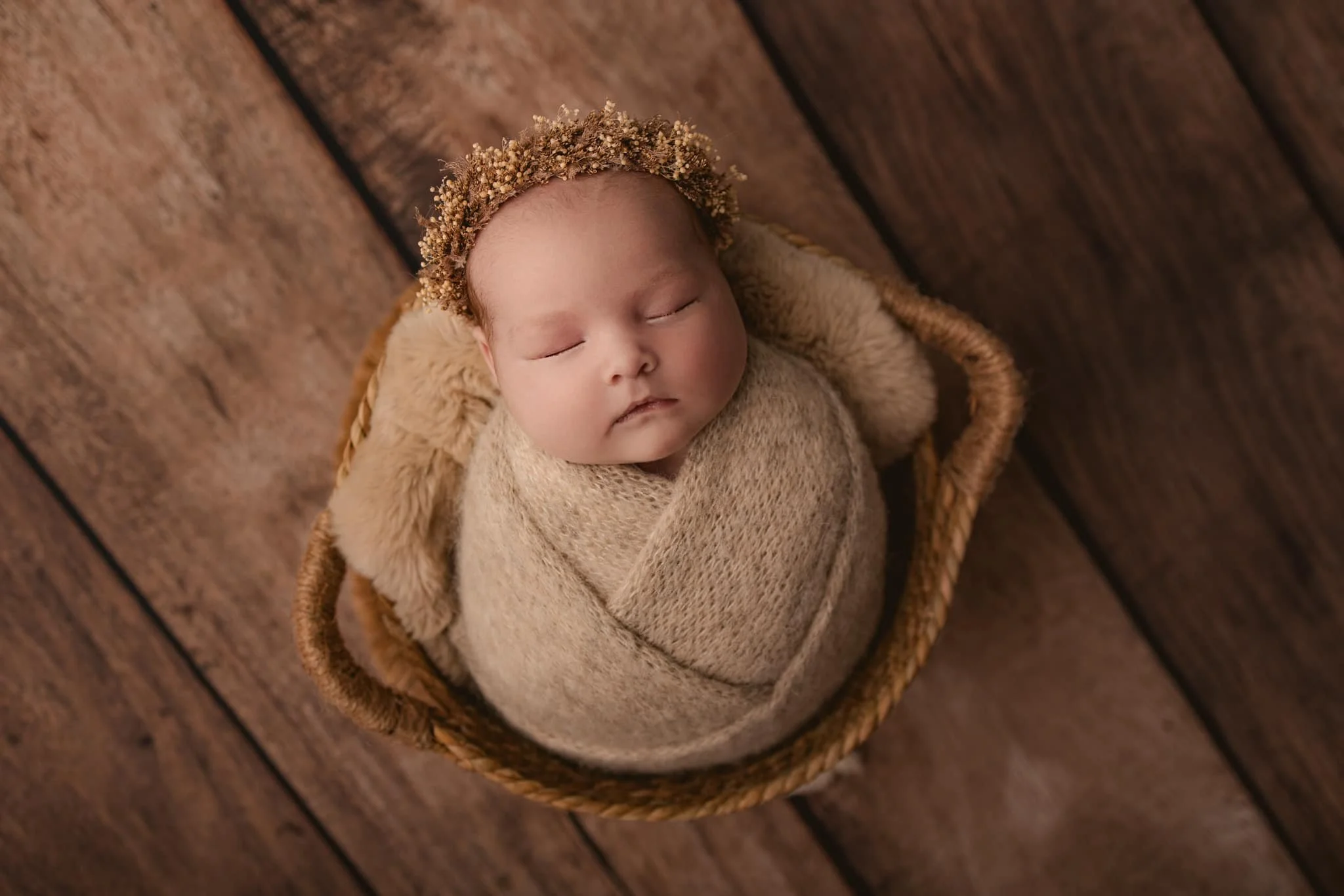 A sleeping baby wrapped in a beige blanket, lying in a small basket on a wooden floor, wearing a floral headband.