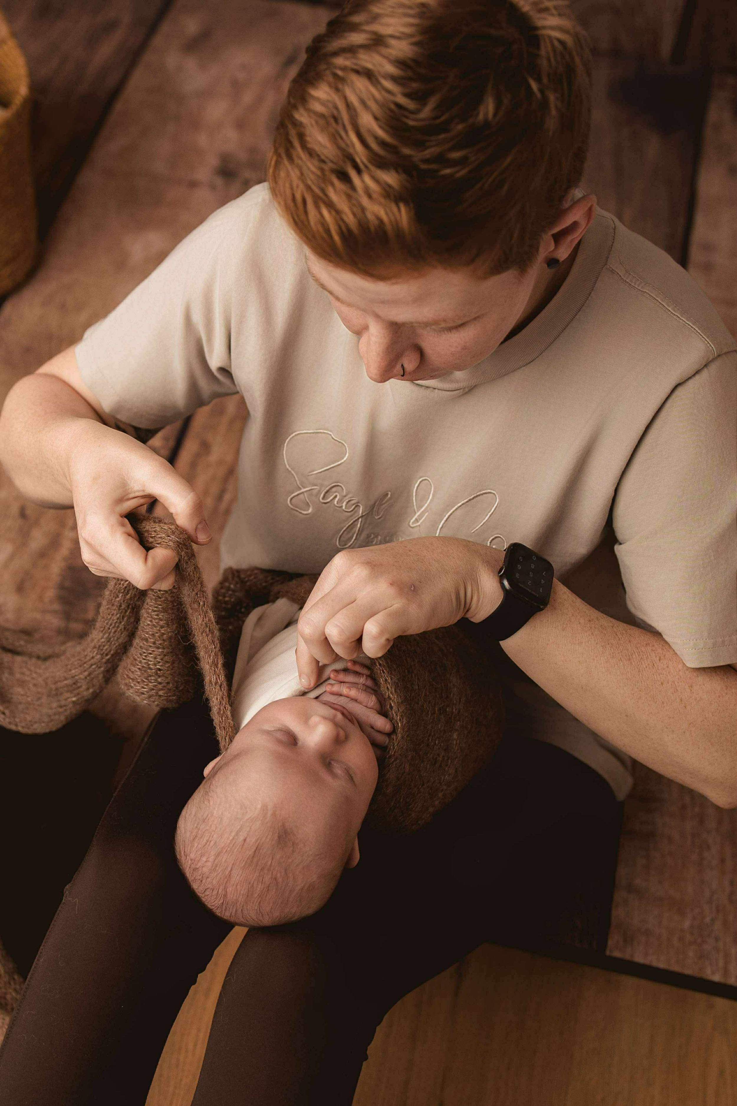 A person with short red hair and a nose piercing gently holds a newborn baby wrapped in a brown knitted blanket, lying on their lap; the person is looking down at the baby and wearing a beige t-shirt and a smartwatch.