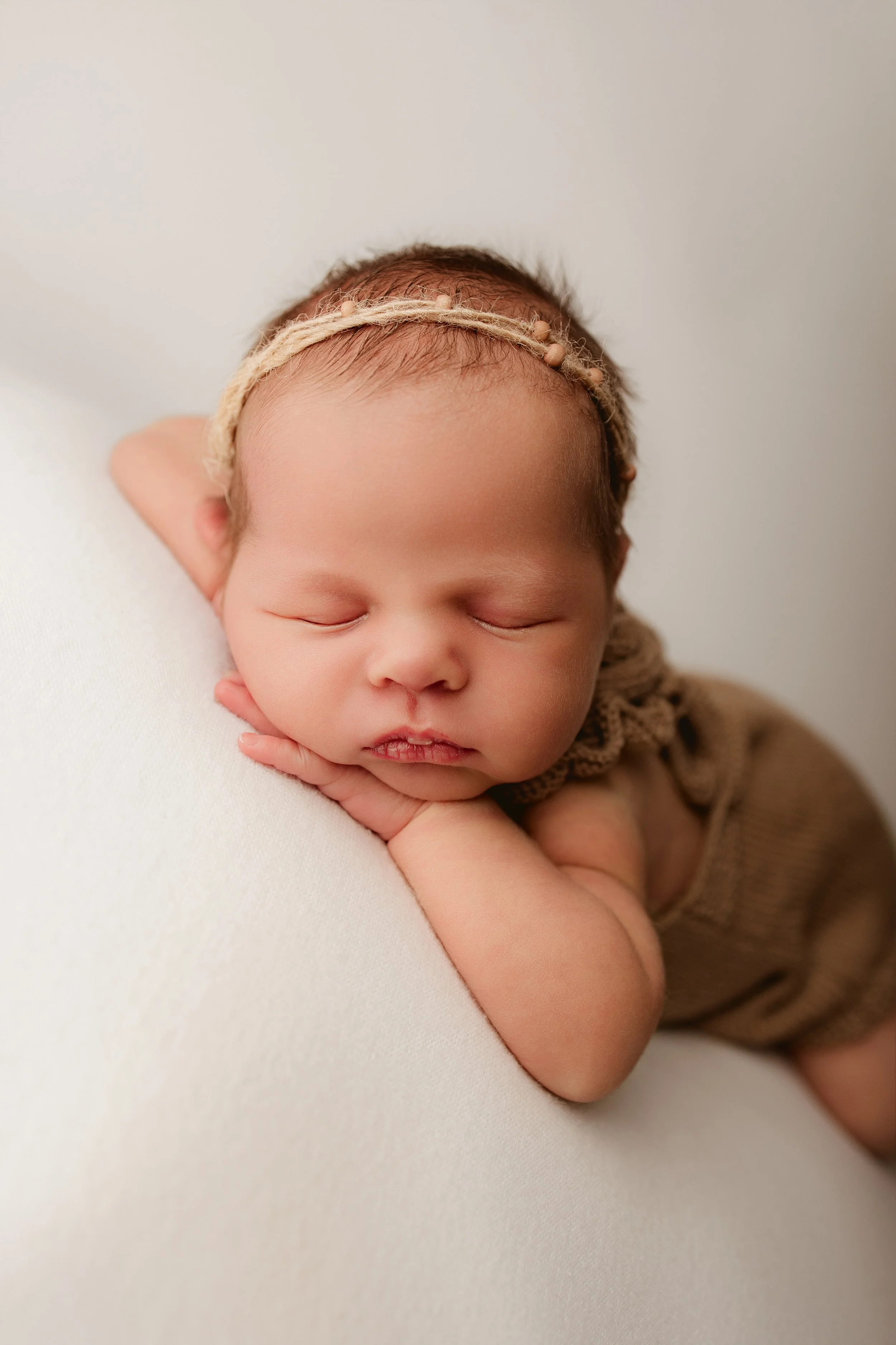 A sleeping newborn baby with a headband, resting their face on their hand on a white surface.