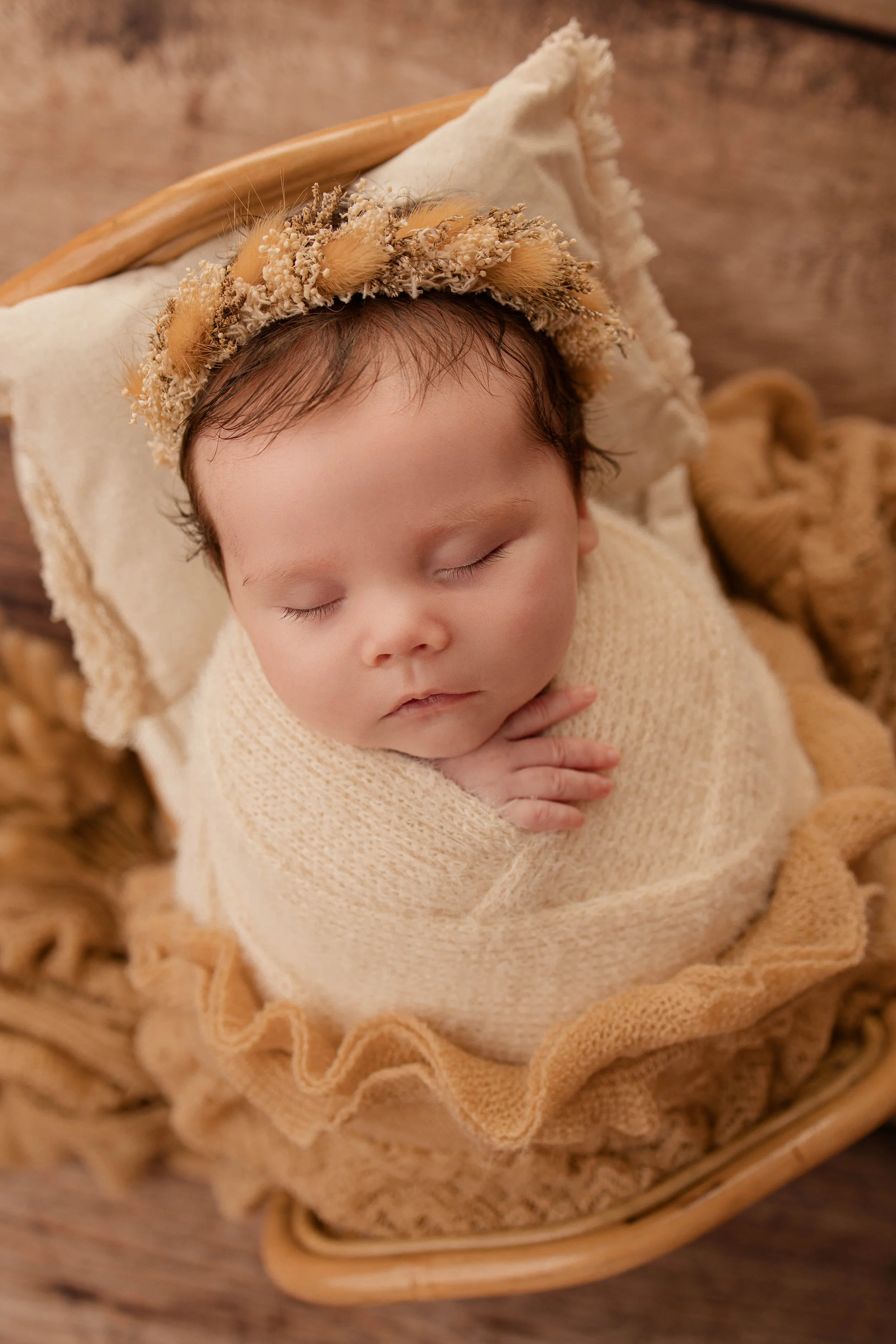 A sleeping baby wrapped in a beige knit blanket, laying in a wicker basket with a ruffled lining, wearing a floral headband with dried flowers.