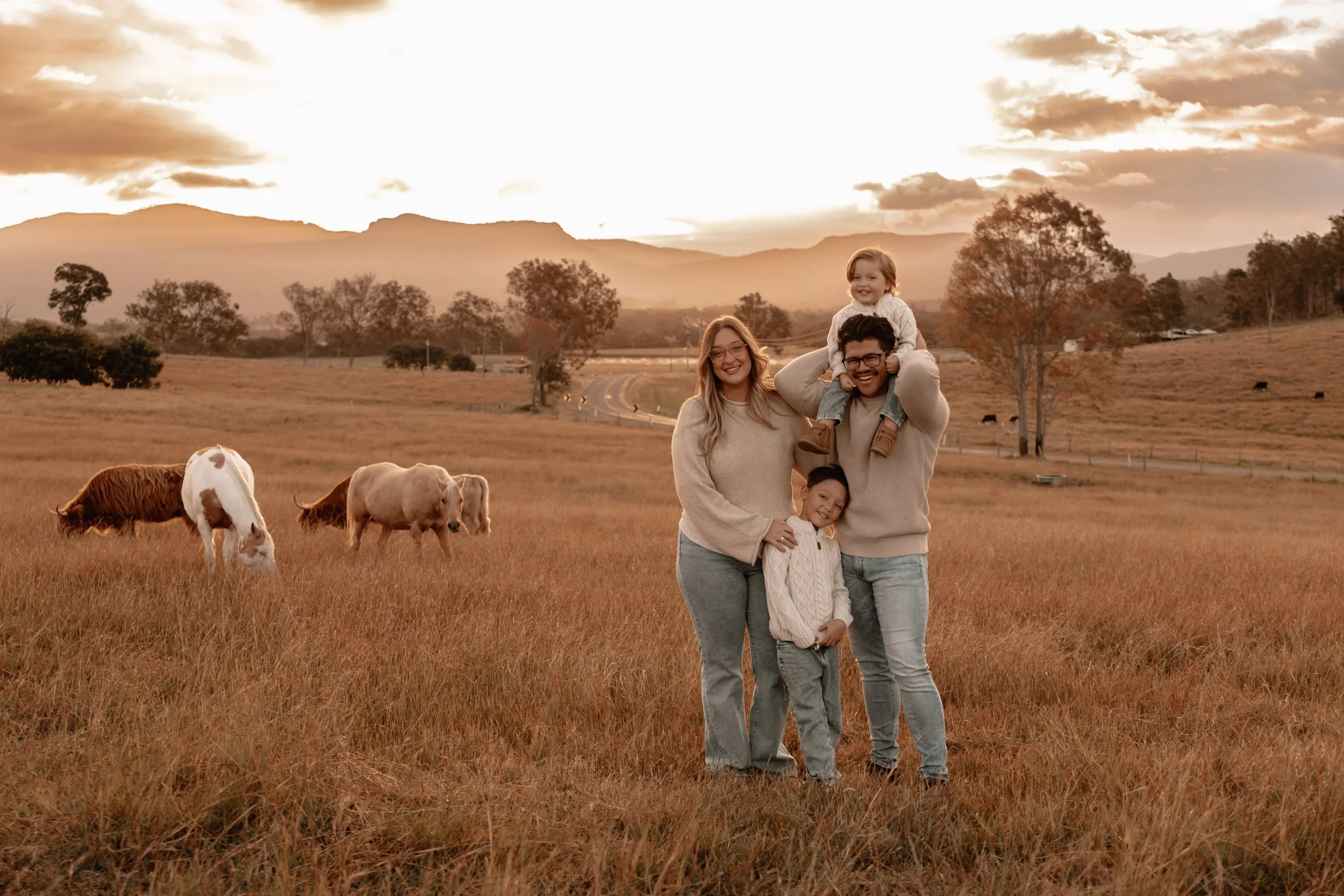 A family of four standing in a field at sunset with mountains and trees in the background, and cows grazing nearby.