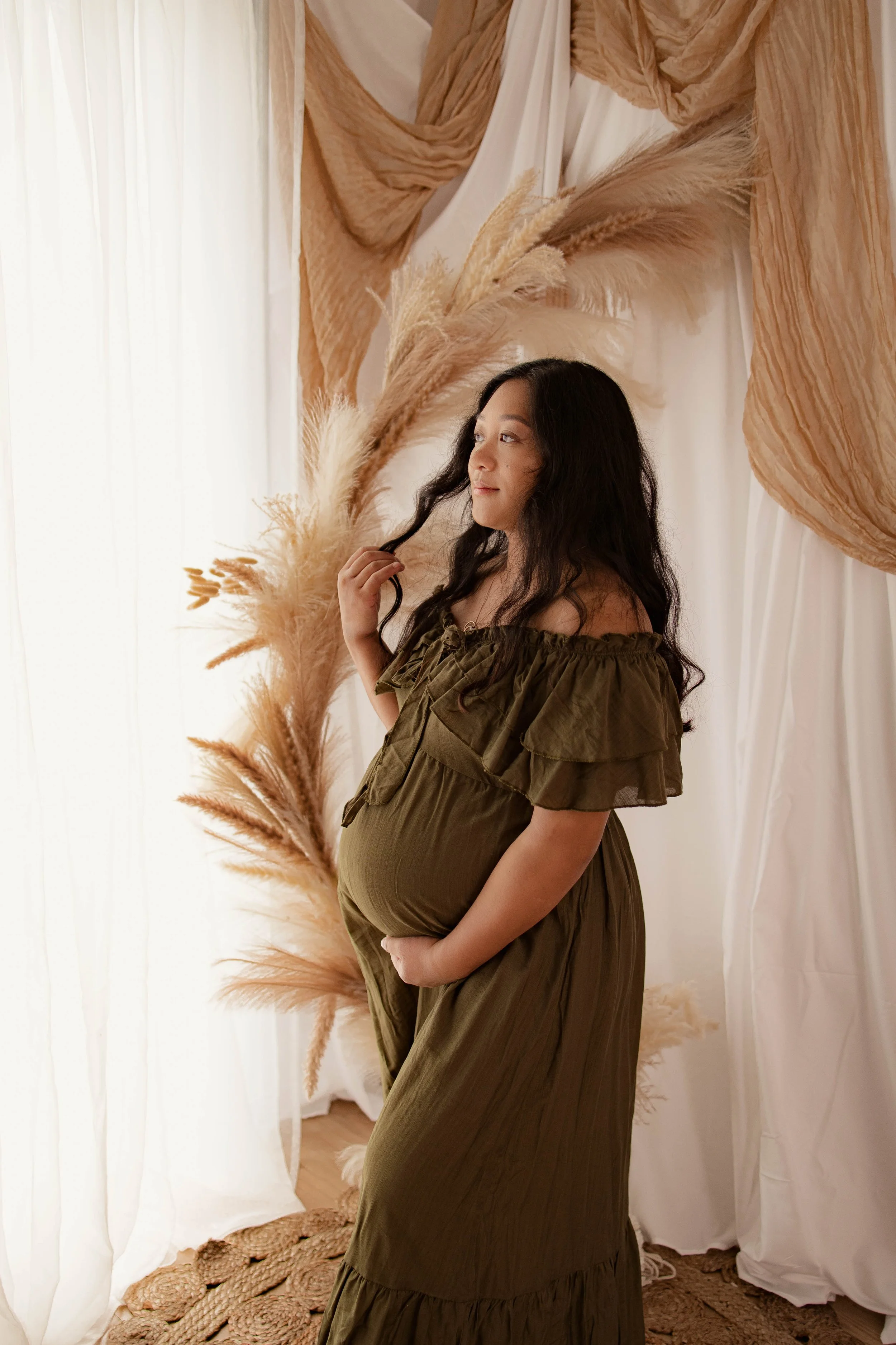 A pregnant woman with long black hair standing in a softly lit studio, touching her hair and cradling her belly, surrounded by beige pampas grass and draped fabrics.