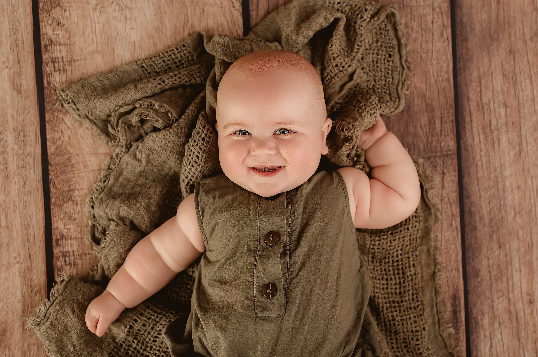 Smiling baby laying on a textured blanket on a wooden floor, wearing a brown sleeveless outfit with buttons.