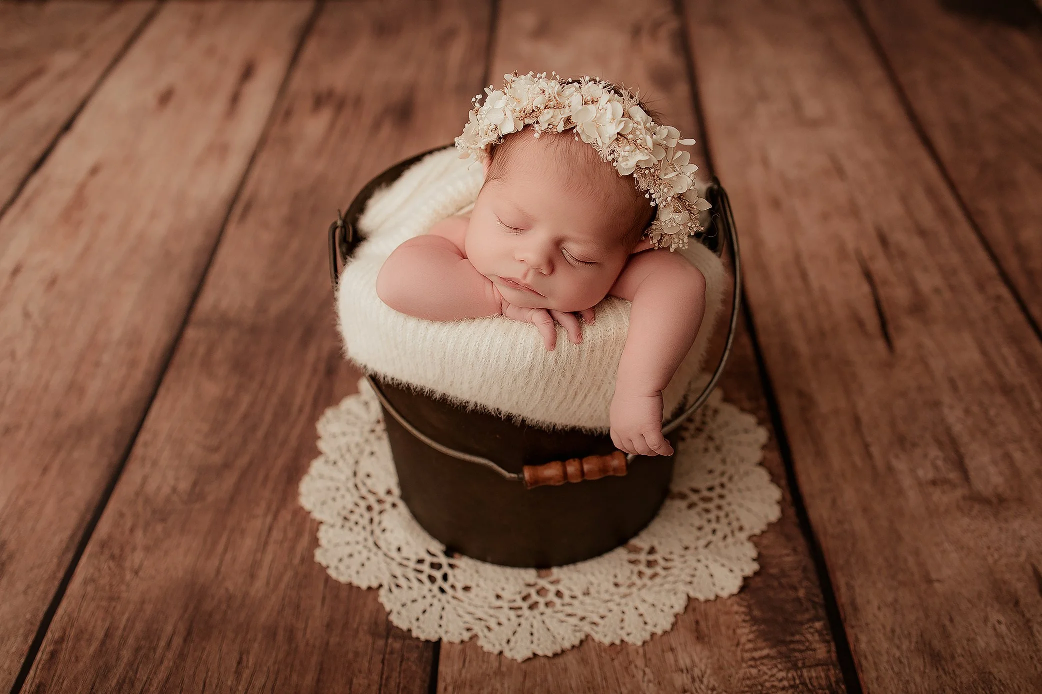 A sleeping newborn baby resting with arms crossed on a knit blanket inside a small black bucket, wearing a flower crown, on a wooden floor with a doily underneath.
