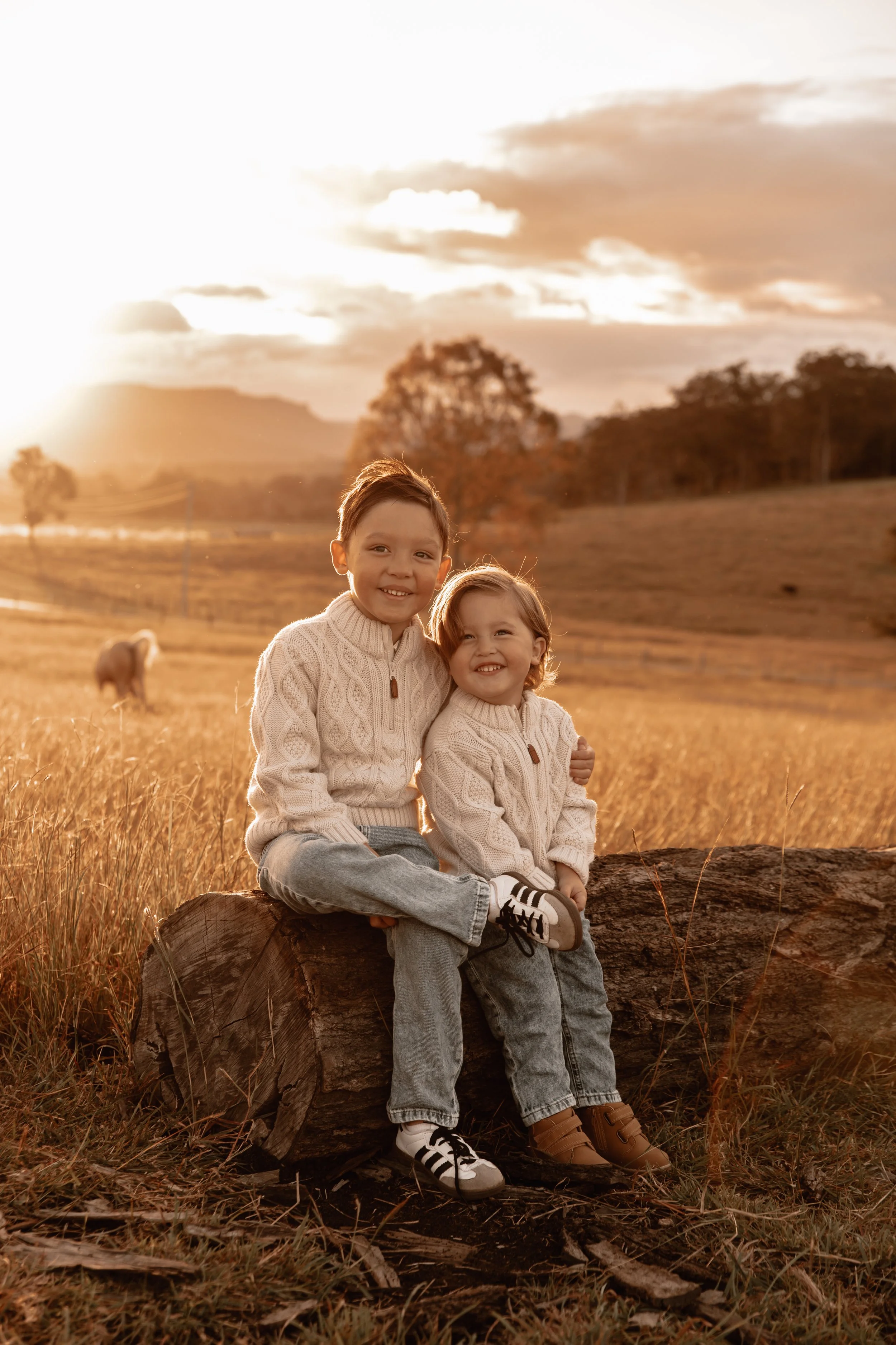 Two young children, a boy and a girl, sitting on a large log in a grassy field during sunset, both wearing cream-colored sweaters, smiling with their arms around each other.