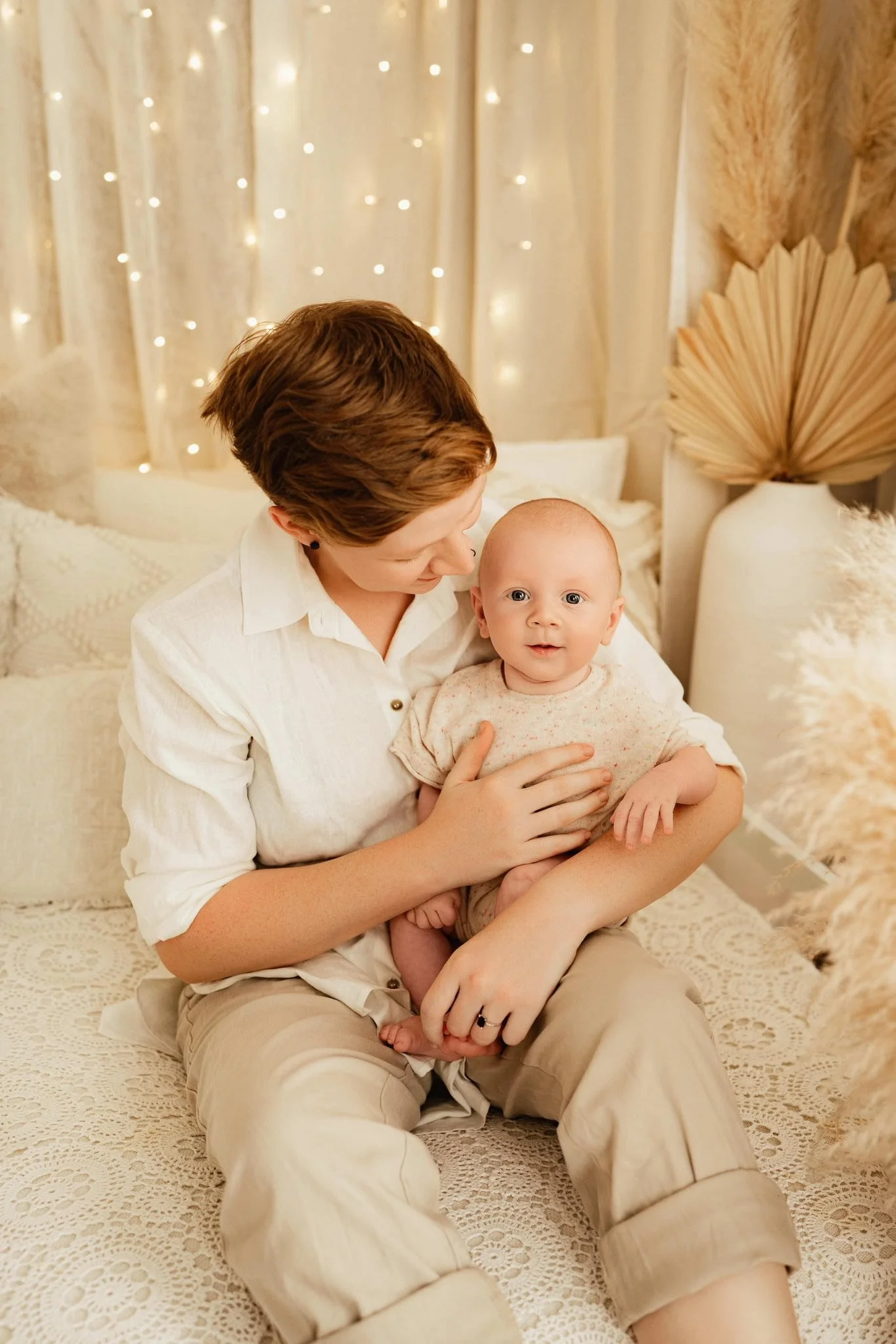 A woman with short brown hair sitting on a lace-covered bed, holding a baby with bright blue eyes and a light-colored onesie, in a warmly lit room decorated with string lights and dried pampas grass.