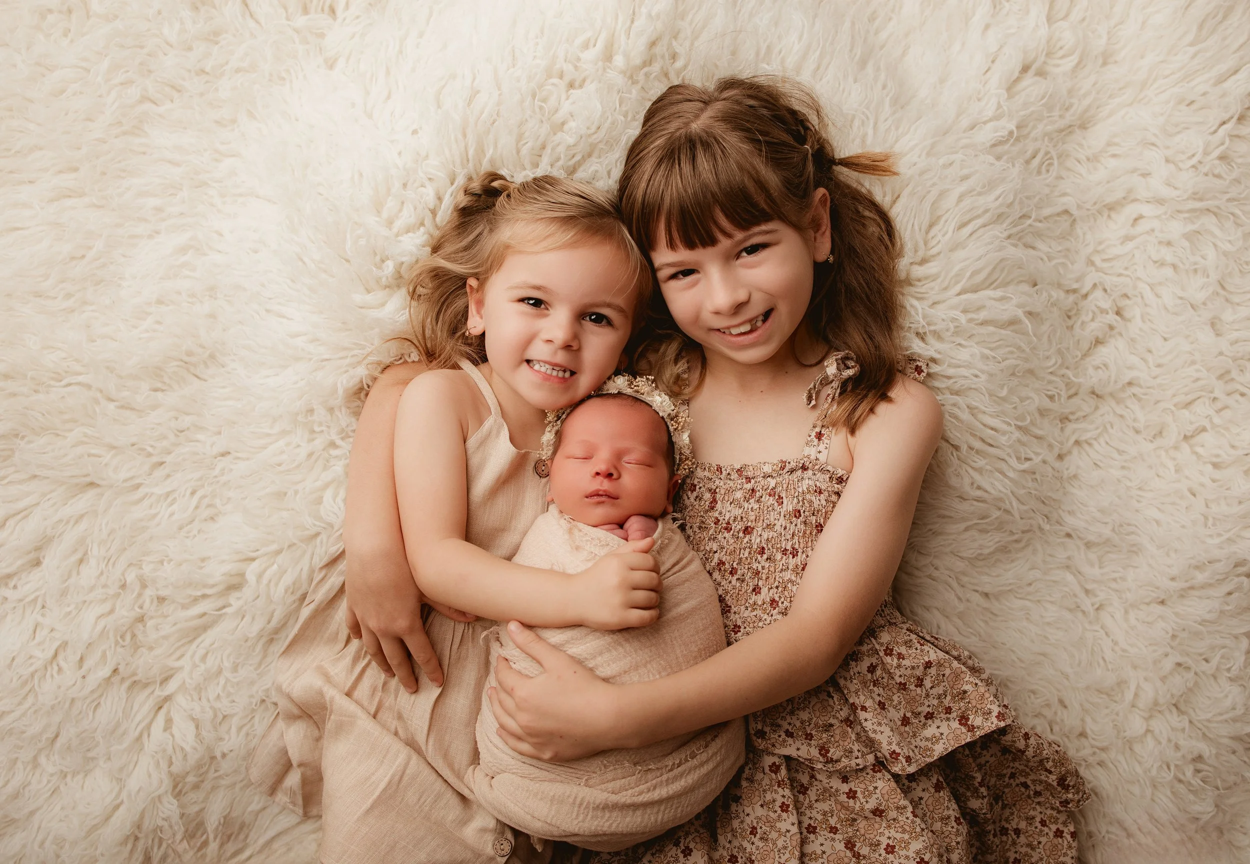 Three young girls, one newborn and two older, lying on a fluffy cream-colored blanket and smiling at the camera.