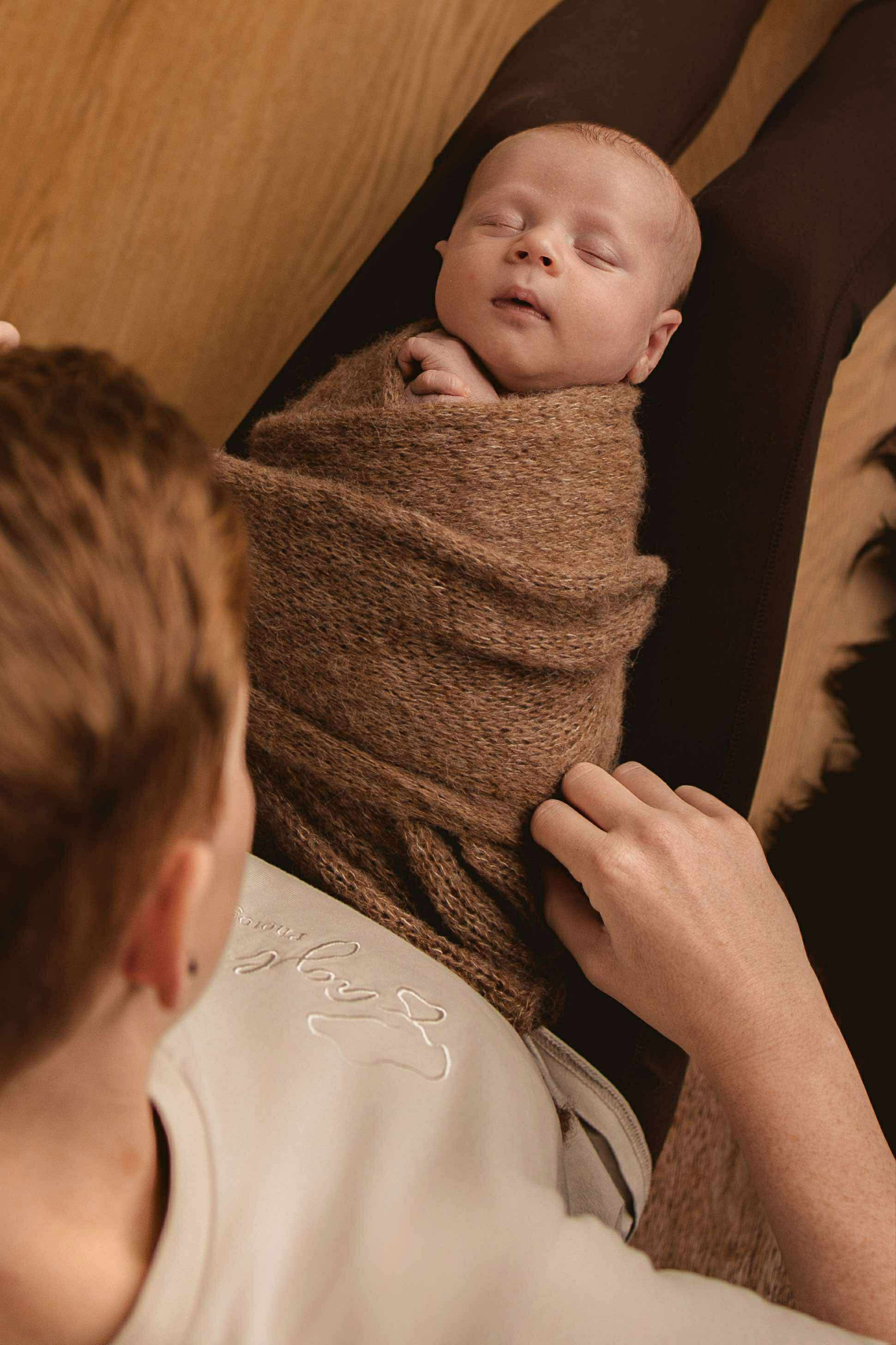 A newborn baby wrapped in a brown blanket lying on a black surface, with an adult woman touching the blanket, and a person with red hair and a tattooed arm looking at the baby.