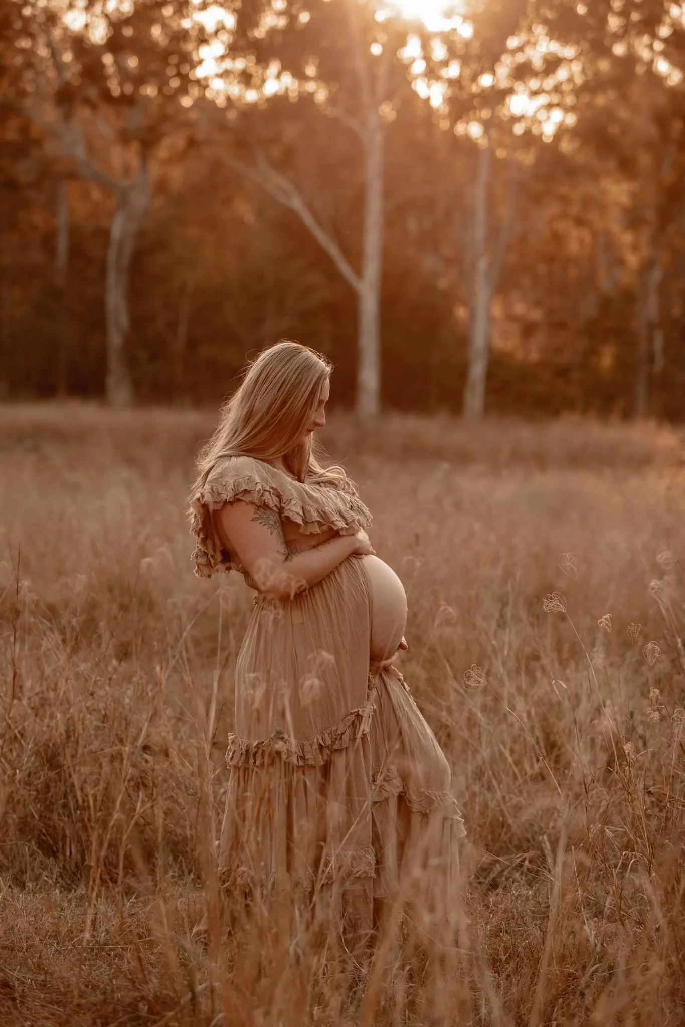 A pregnant woman with long blonde hair stands in a grassy field during sunset, cradling her belly gently and looking down.