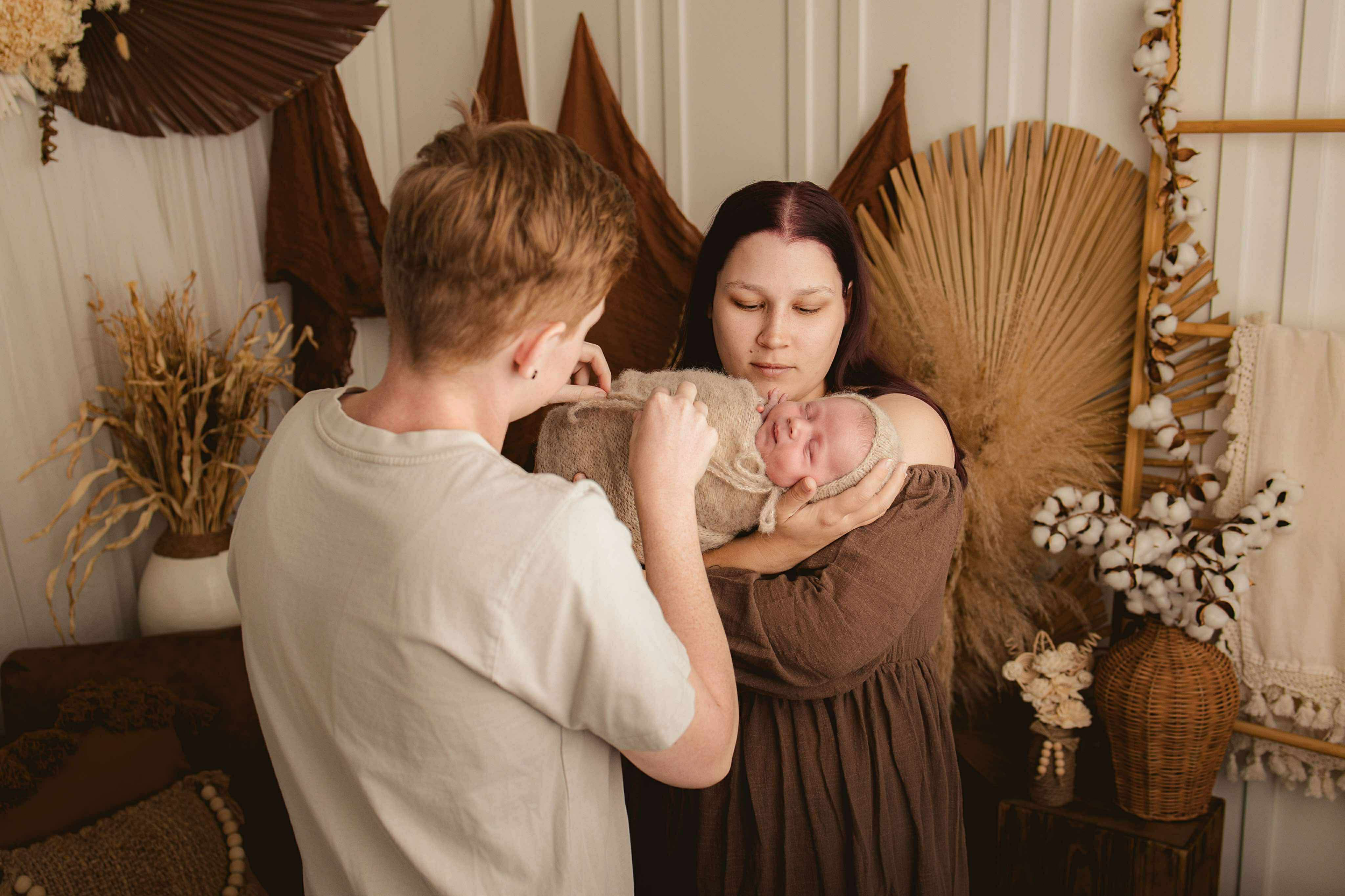 A woman holding a newborn baby, with a man touching the baby's hand. The setting has warm decorations including dried plants and woven art on the wall.