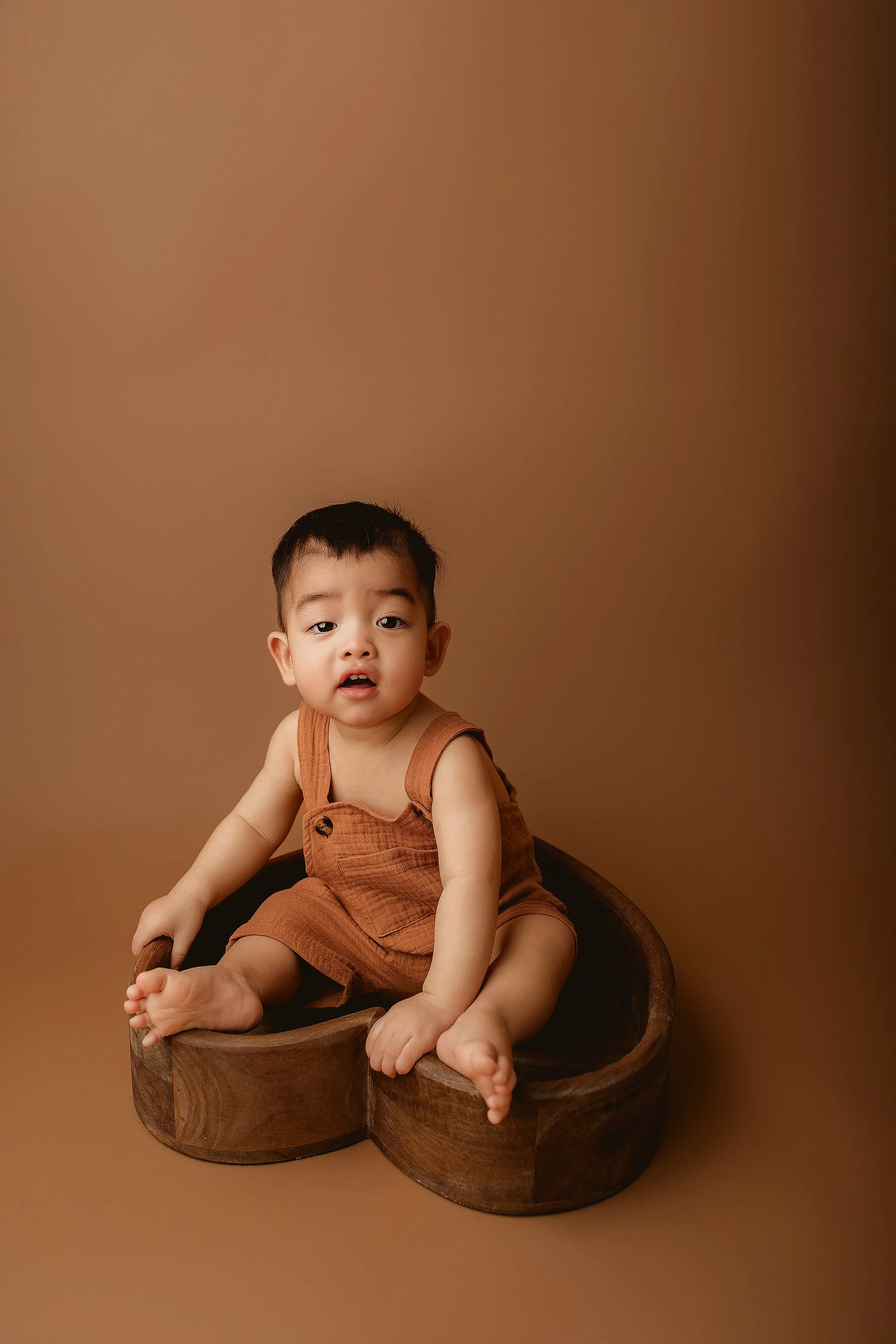 Young boy sitting in a round wooden bowl, wearing a brown romper, against a brown gradient background.