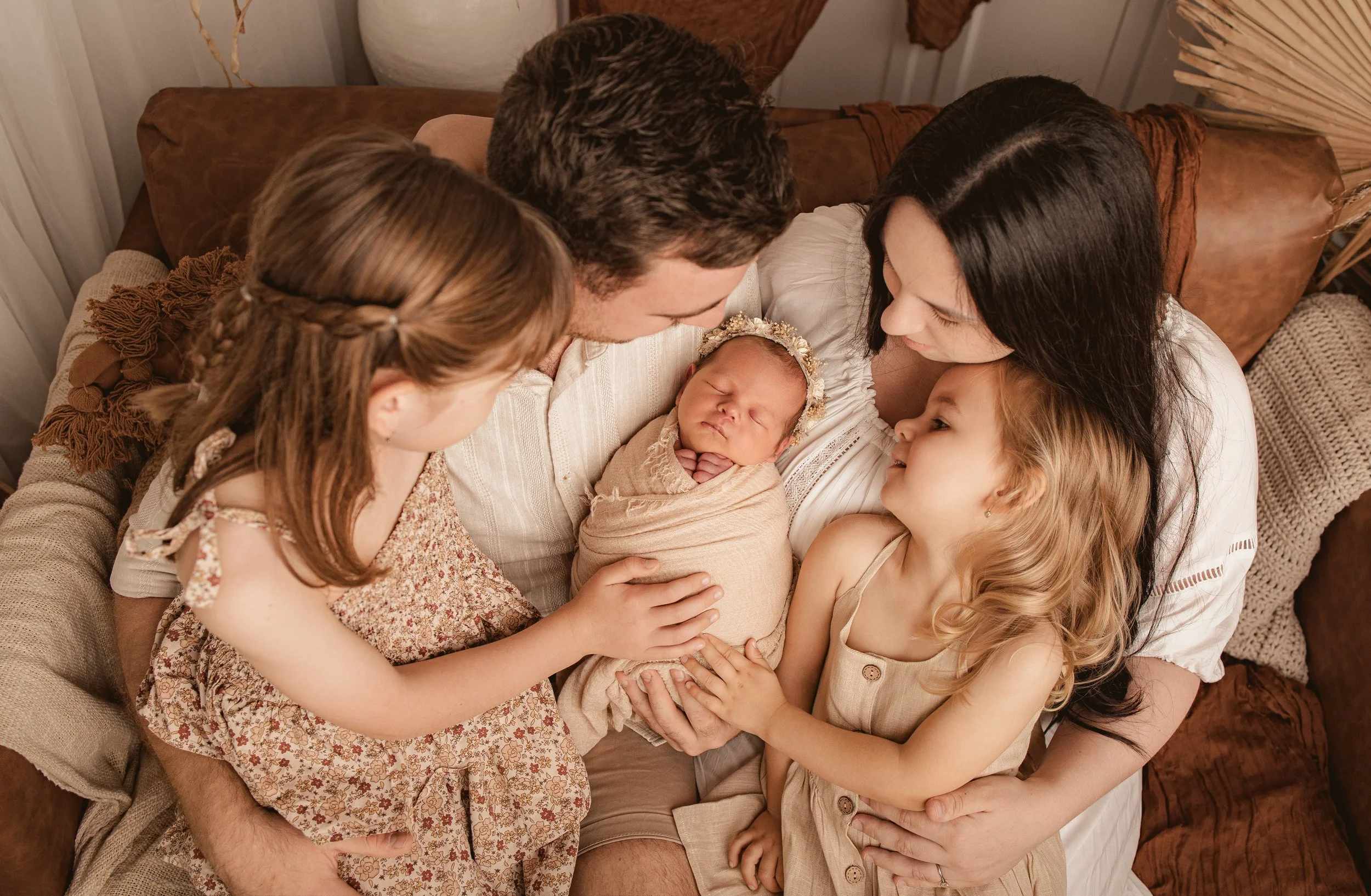 A family of five celebrating a newborn baby girl, with parents, two young daughters, and the baby girl all gathered on a sofa.