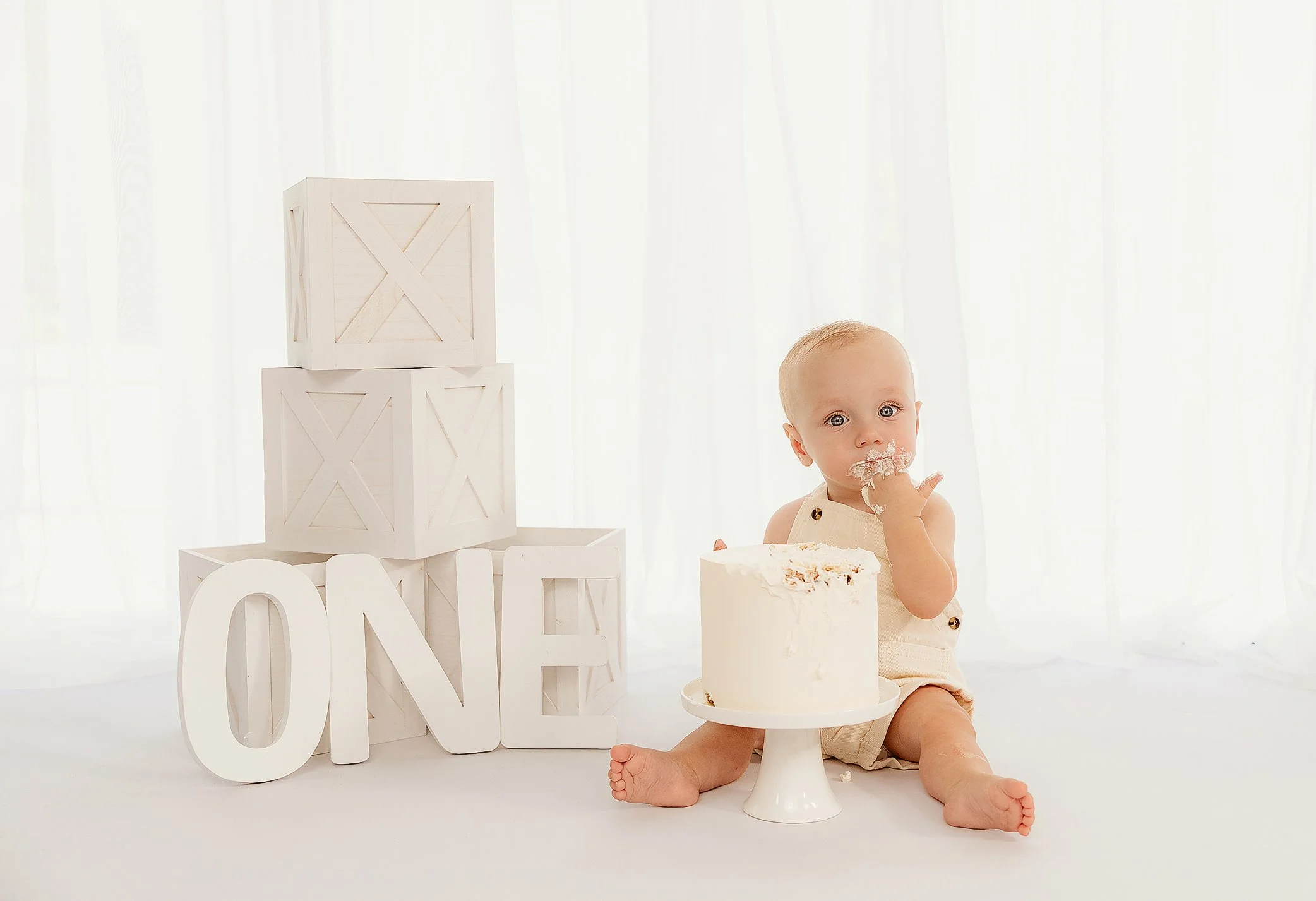 Baby sitting on white floor with a partially eaten cake in front, surrounded by white decorative blocks spelling 'ONE', indicating a first birthday celebration, with a plain white/cream background.