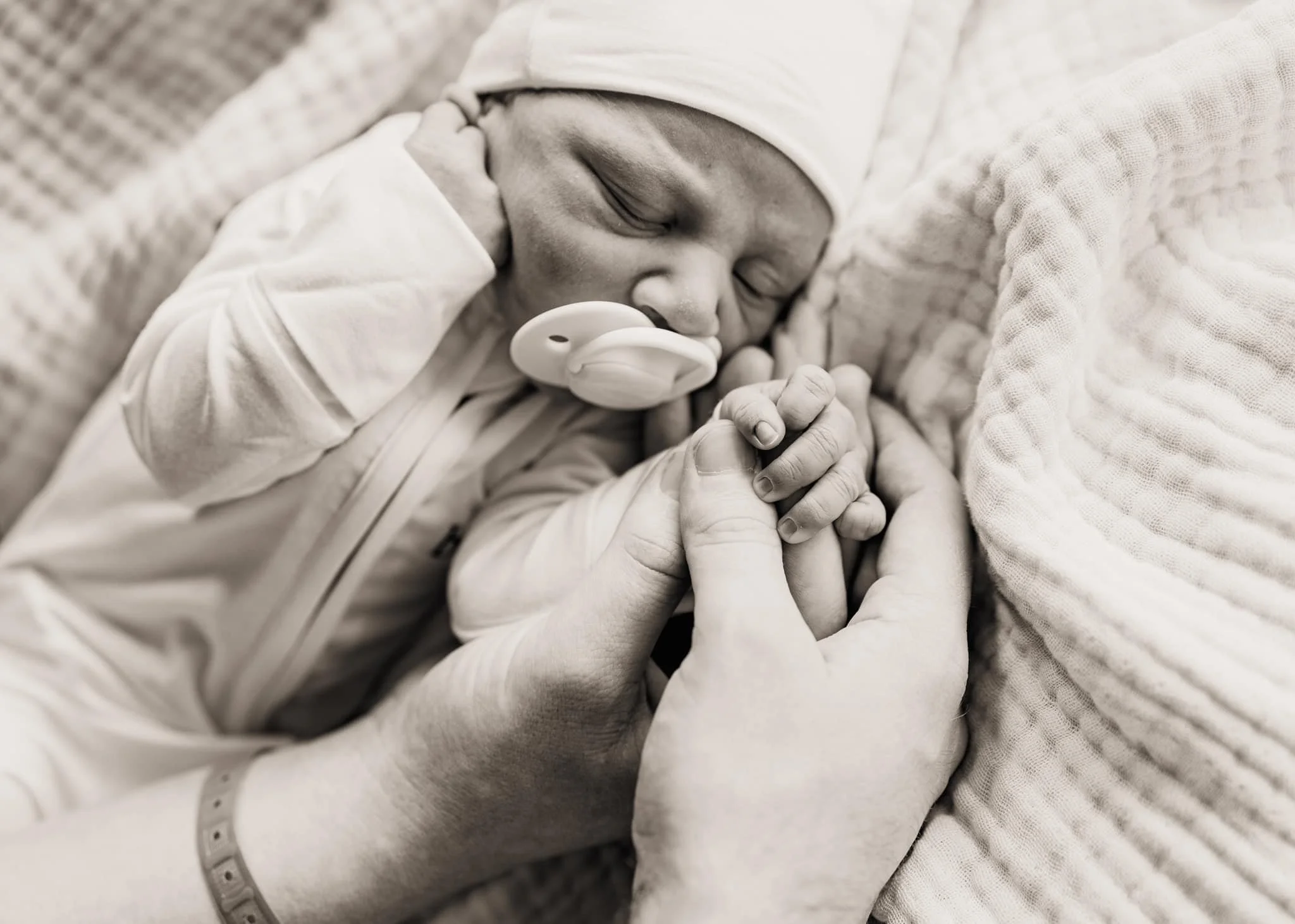 A newborn baby with a pacifier in mouth, resting on a pillow, holding an adult's finger, with a hospital wristband on the adult's wrist.