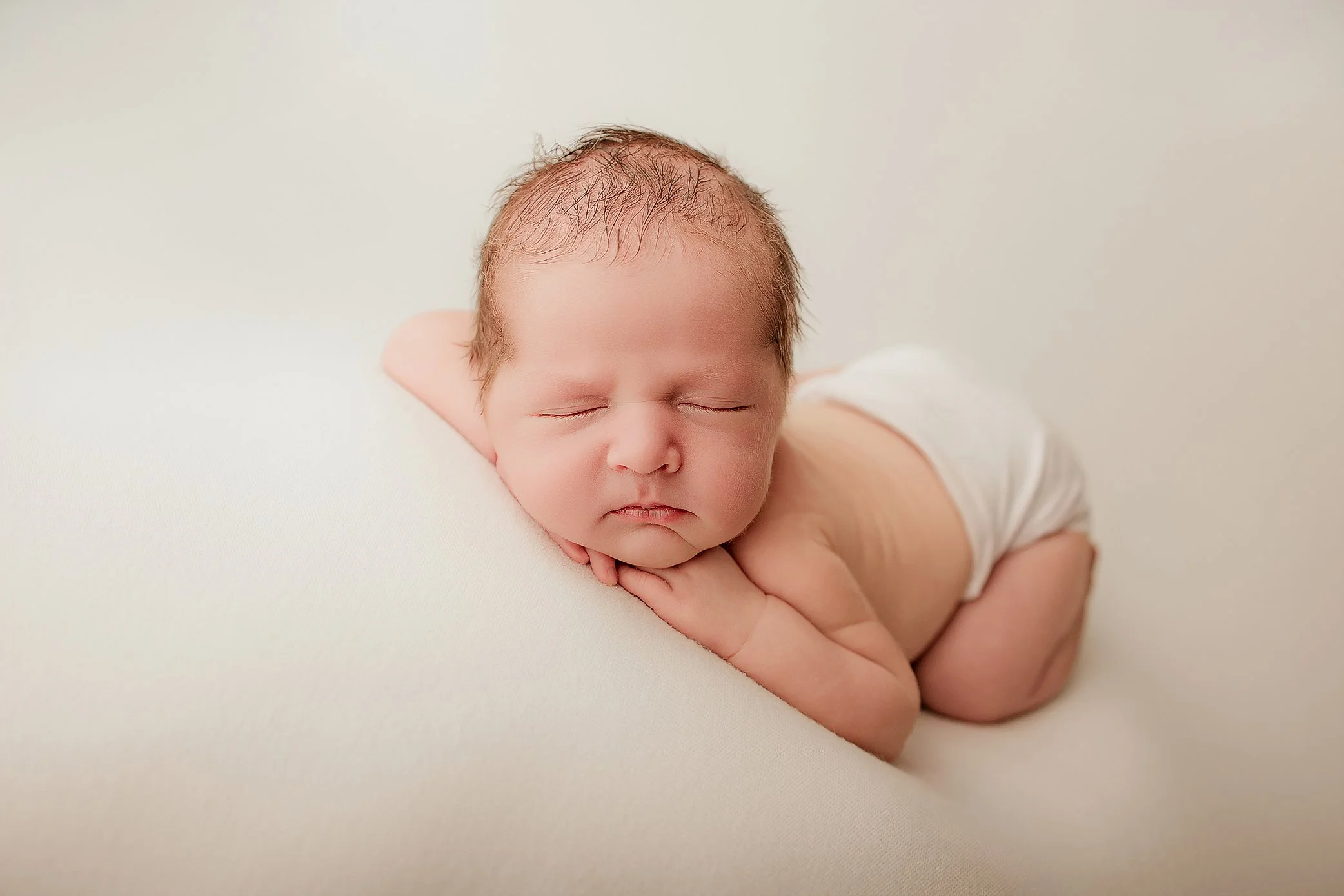 A sleeping newborn baby lying on a white surface, resting its head on its hands, wearing only a diaper.