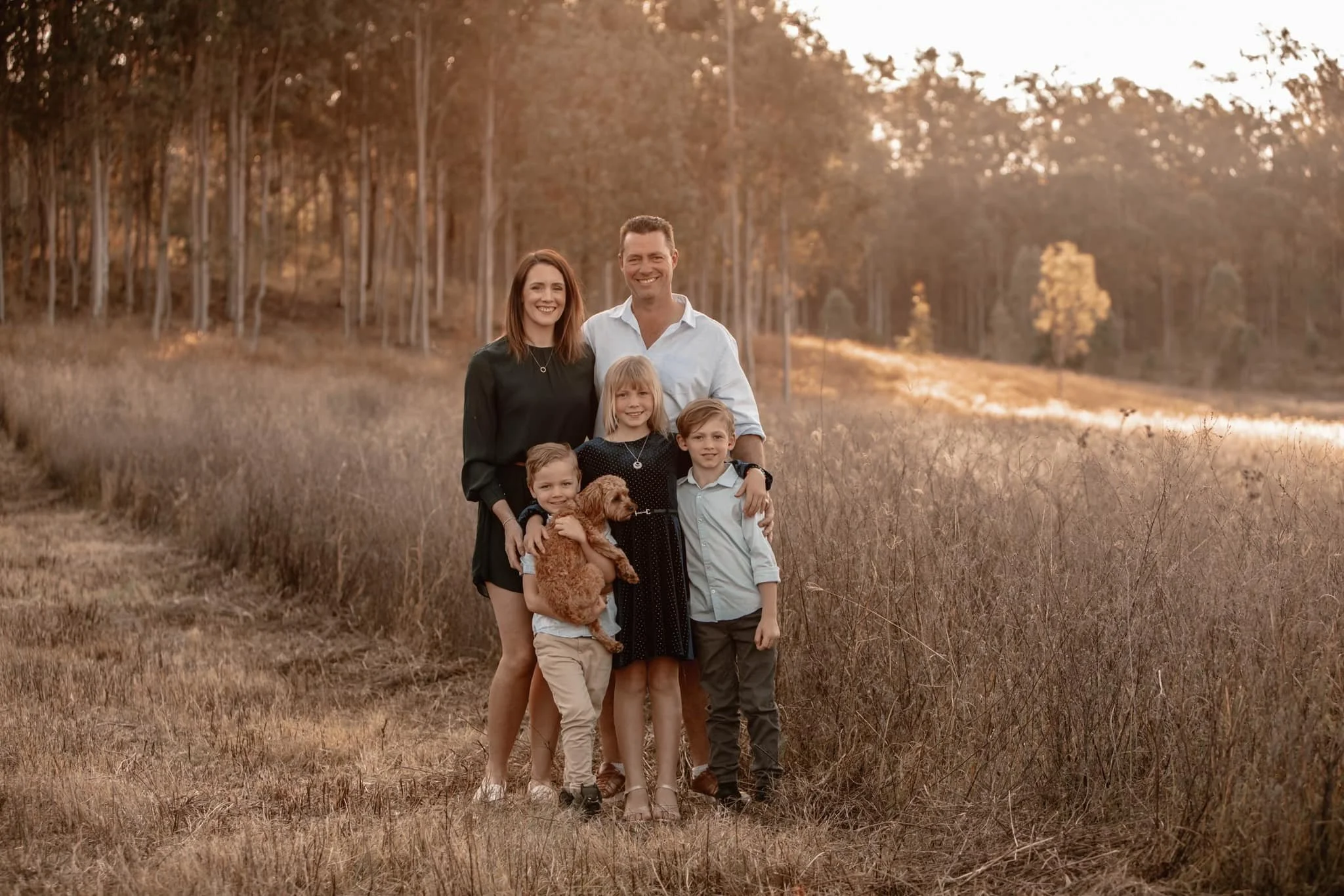 A family of six, including a dog, standing together outdoors in a grassy field during sunset.