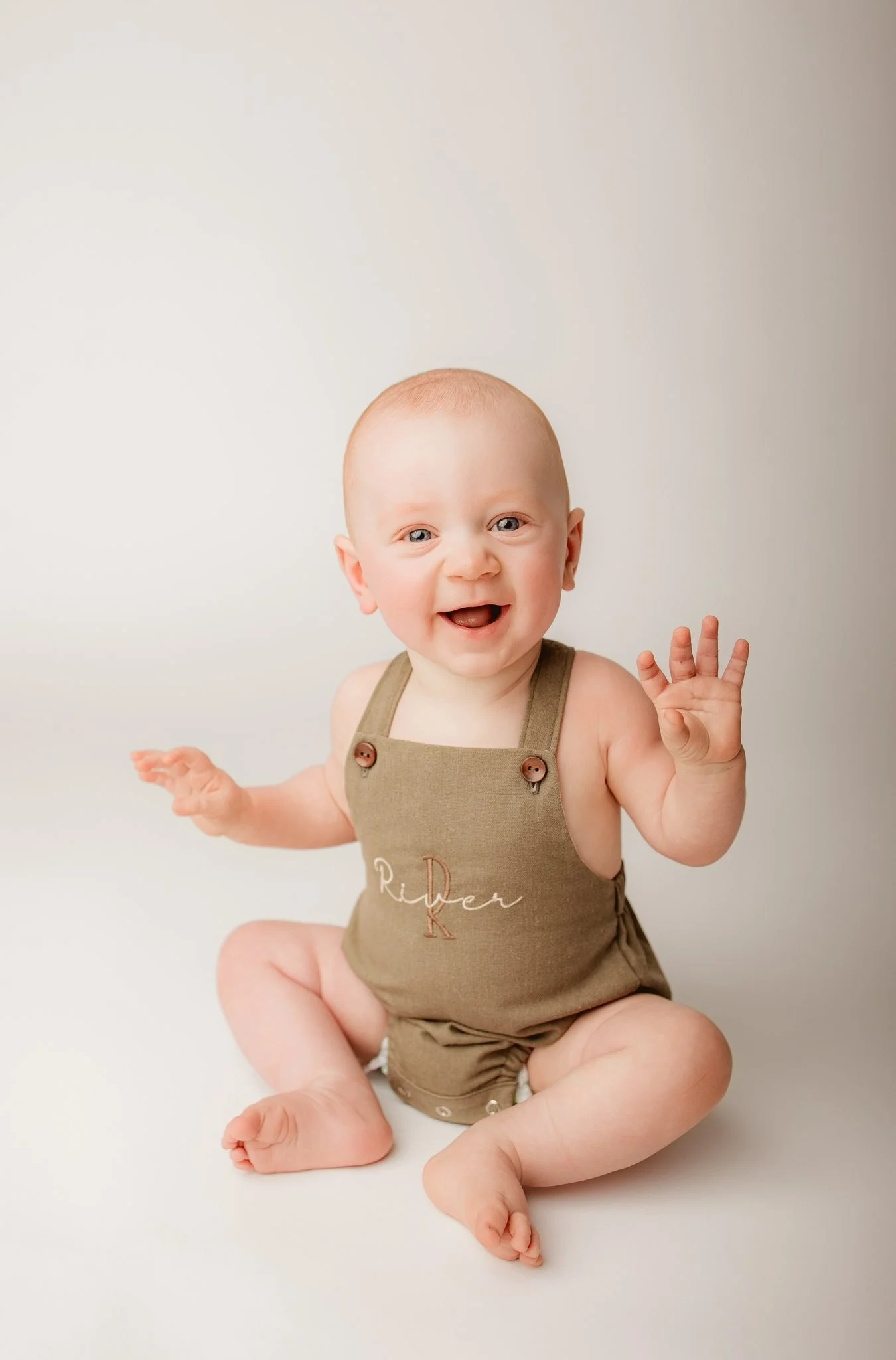 A smiling baby sitting on a white surface, wearing a brown outfit with the text 'River R' embroidered on it, waving one hand.