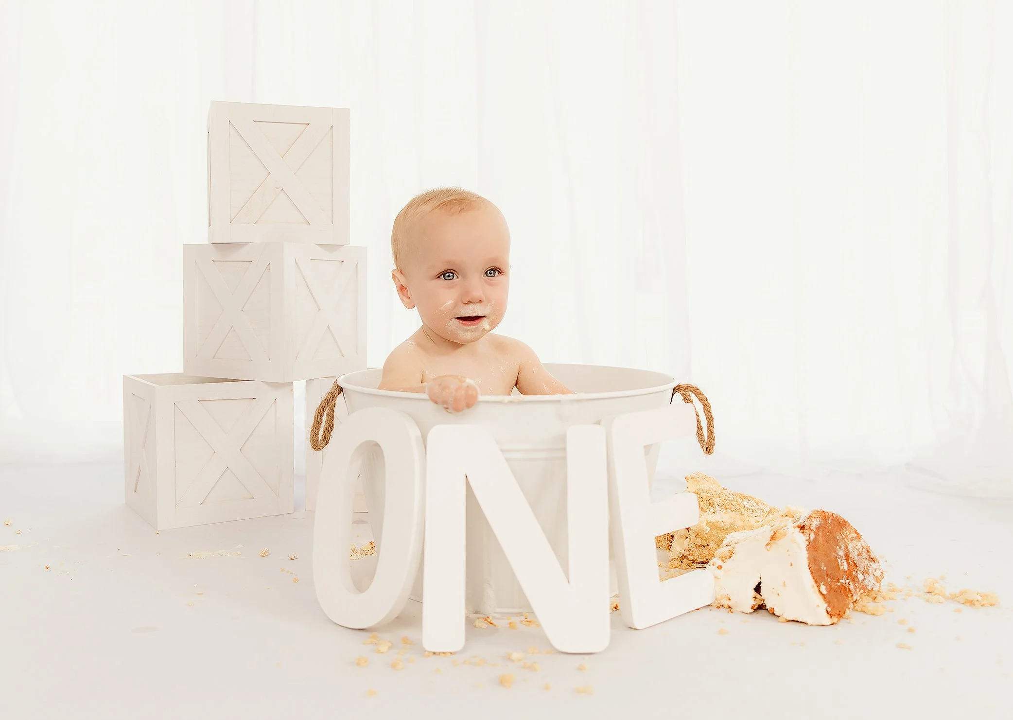 A baby sitting in a white tub marked for a first birthday, surrounded by broken cake and large decorative white letters spelling 'ONE', with white storage cubes in the background and sheer white curtains.