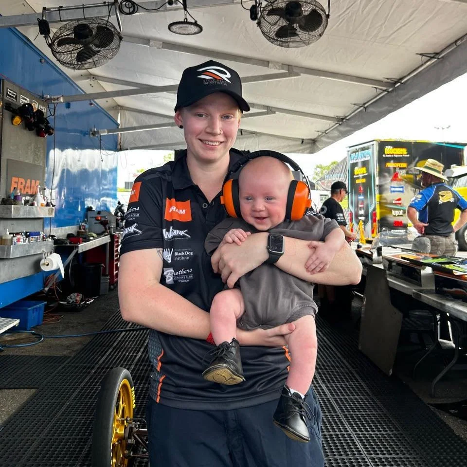 A woman in racing team apparel holding a smiling baby with headphones in a garage or pit area, with racing trailers and team members in the background.