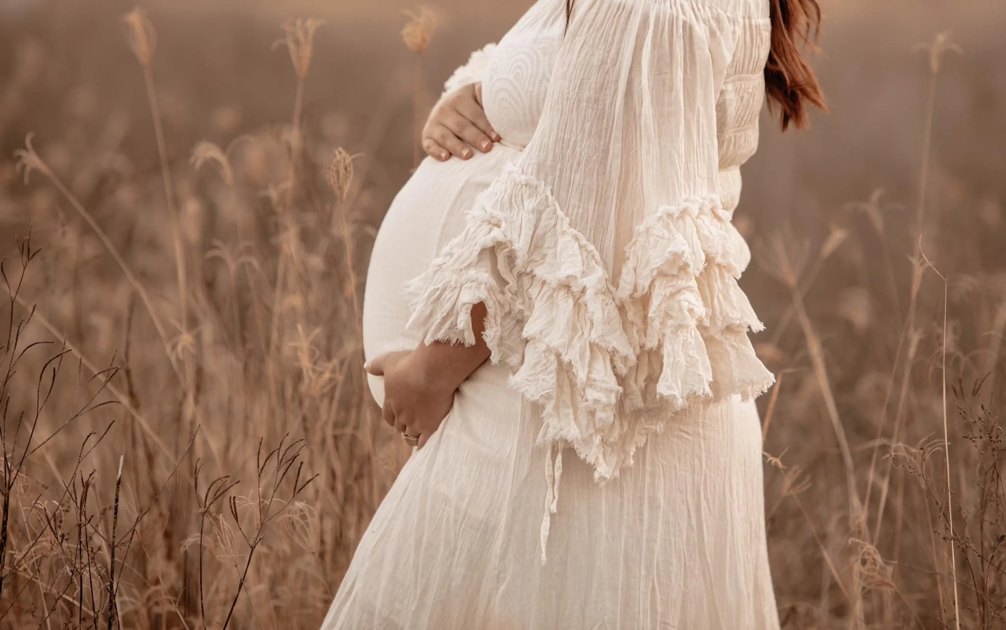 Pregnant woman holding her belly in a field of tall, dried grass, wearing a flowing cream-colored dress with ruffled sleeves.