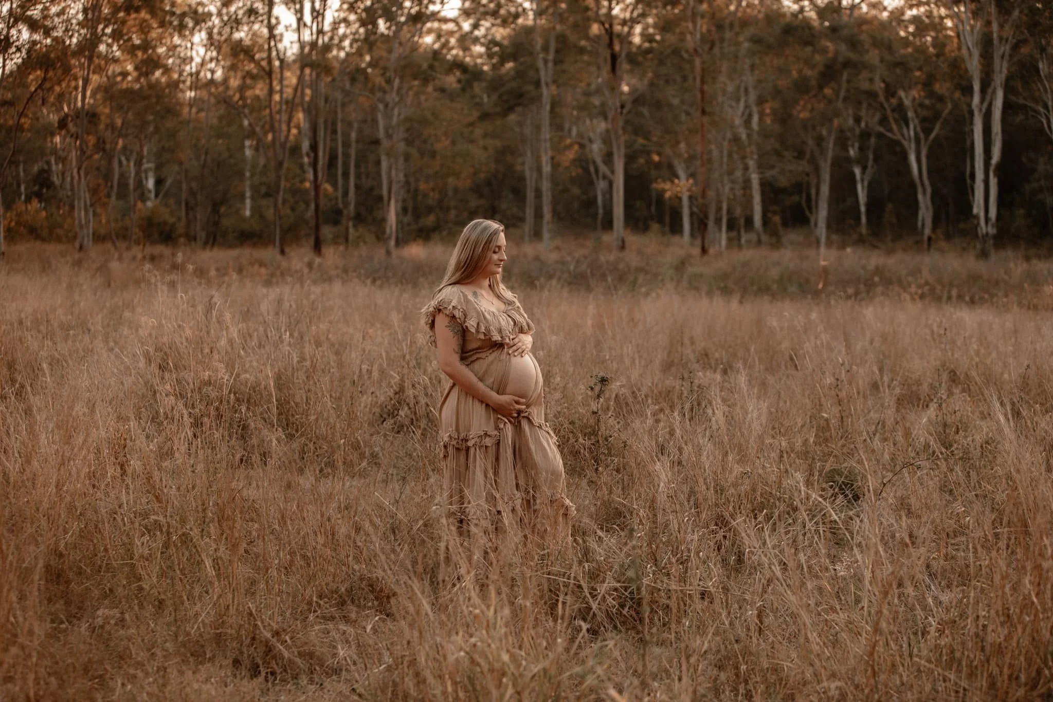 A pregnant woman standing in a field of tall, brown grass with a line of trees in the background during sunset.