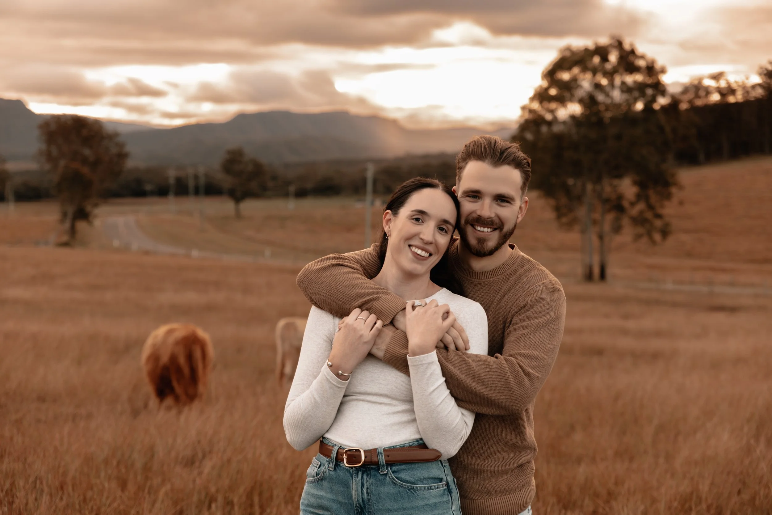 A couple stands close together in a field during sunset, smiling. The man has his arms around the woman, and the woman is holding his hands. The background features trees, mountains, and a cloudy sky, with a couple of cows grazing nearby.