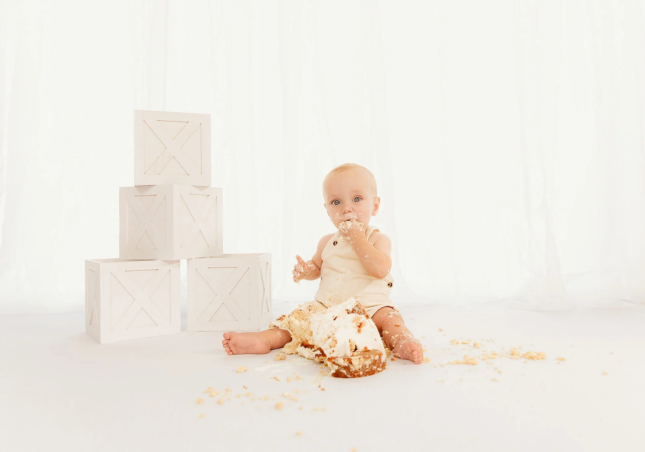 A baby sitting on the floor surrounded by cake, with some cake on their face and hands, and white wooden crates stacked in a pyramid nearby, in a bright, white setting.